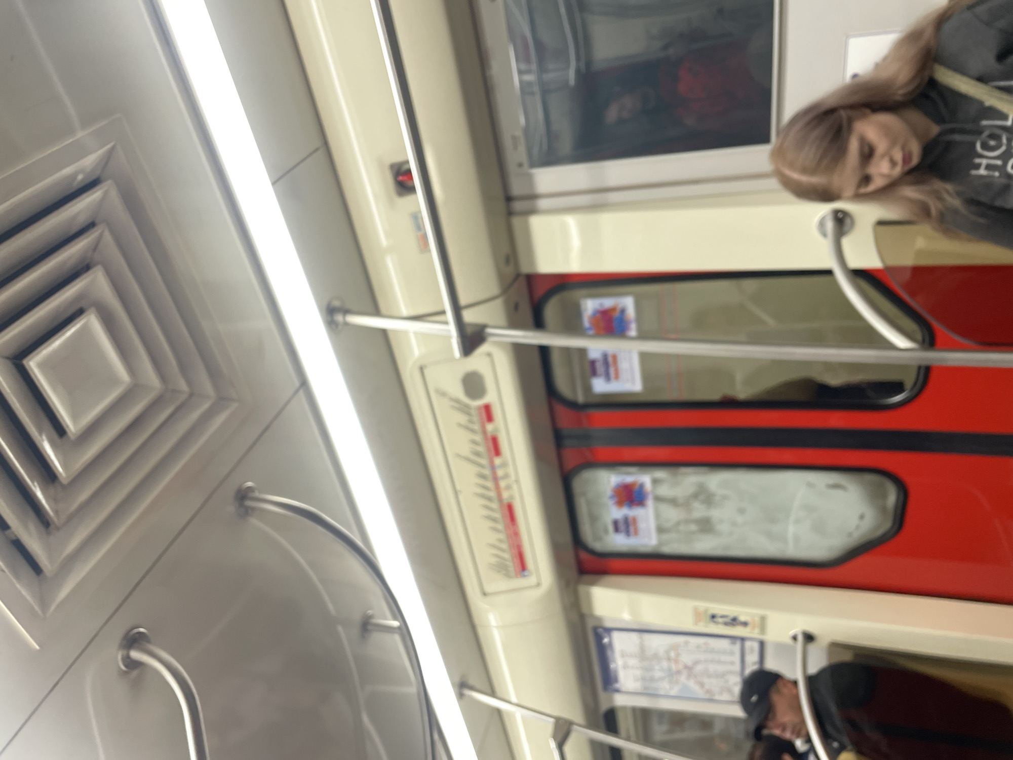 The image captures the interior of a subway car in Saint Petersburg, Russia, viewed from an oblique, slightly elevated angle. The car features a cream-colored ceiling and walls, brightly illuminated by a long, linear fluorescent light fixture running along the ceiling. A square, layered air vent is visible on the left side of the ceiling.
Chrome grab rails are prominent, extending both horizontally and vertically across the car, providing support for passengers. The train's windows are distinctively framed in bright red, contrasting with the lighter interior.
On the right side of the frame, a young woman with long, light brown hair is visible, resting her head on a surface, suggesting she might be asleep or tired. She is wearing a dark grey or black hoodie, with the letters "HOL" partially visible on the fabric. A blurred reflection or another person in a red garment can be seen in the upper window above her.
Further down the carriage, a vertical panel displaying a series of numbers and red lines, likely a station indicator or route progression, is mounted on the wall. Below this, a standard subway line map is affixed, though the text is unreadable due to blur. Another person, wearing a dark cap, is faintly visible in a lower window reflection or through the glass. The scene depicts typical daily commute activity within a modern metro environment.