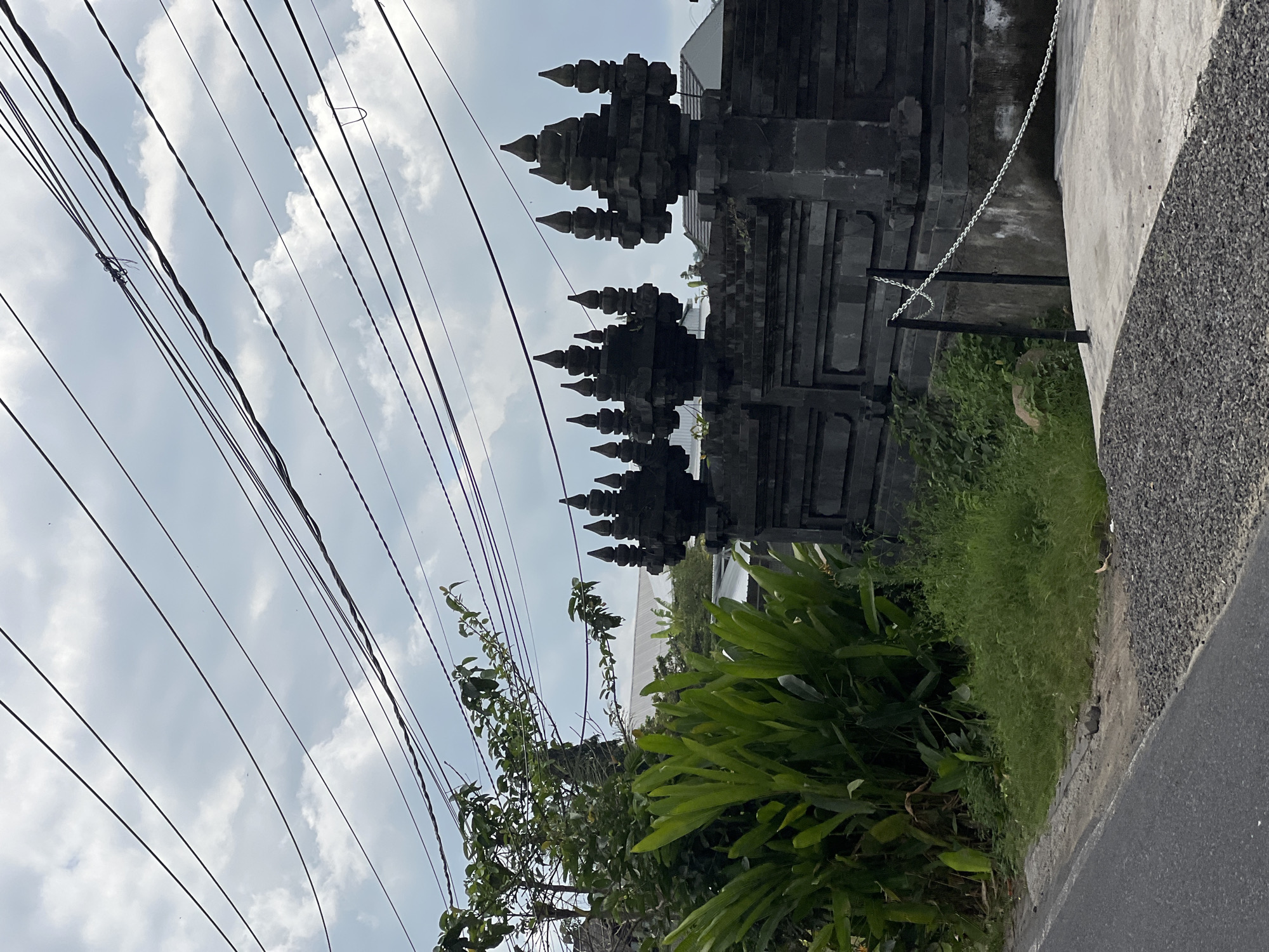 A photograph captures a street view in Abiansemal, Indonesia. The foreground shows a paved road on the left, transitioning to a gravel shoulder and then a concrete walkway. Adjacent to the walkway is a dark, ornate stone wall, characteristic of Balinese architecture, with intricate tiered finials along its top. To the left of the wall, lush green vegetation, including large-leafed plants and a tree, thrives. Overhead, a dense network of dark electrical wires stretches across the cloudy sky, creating a stark contrast with the natural and architectural elements below. The scene is devoid of people or any discernible activity.