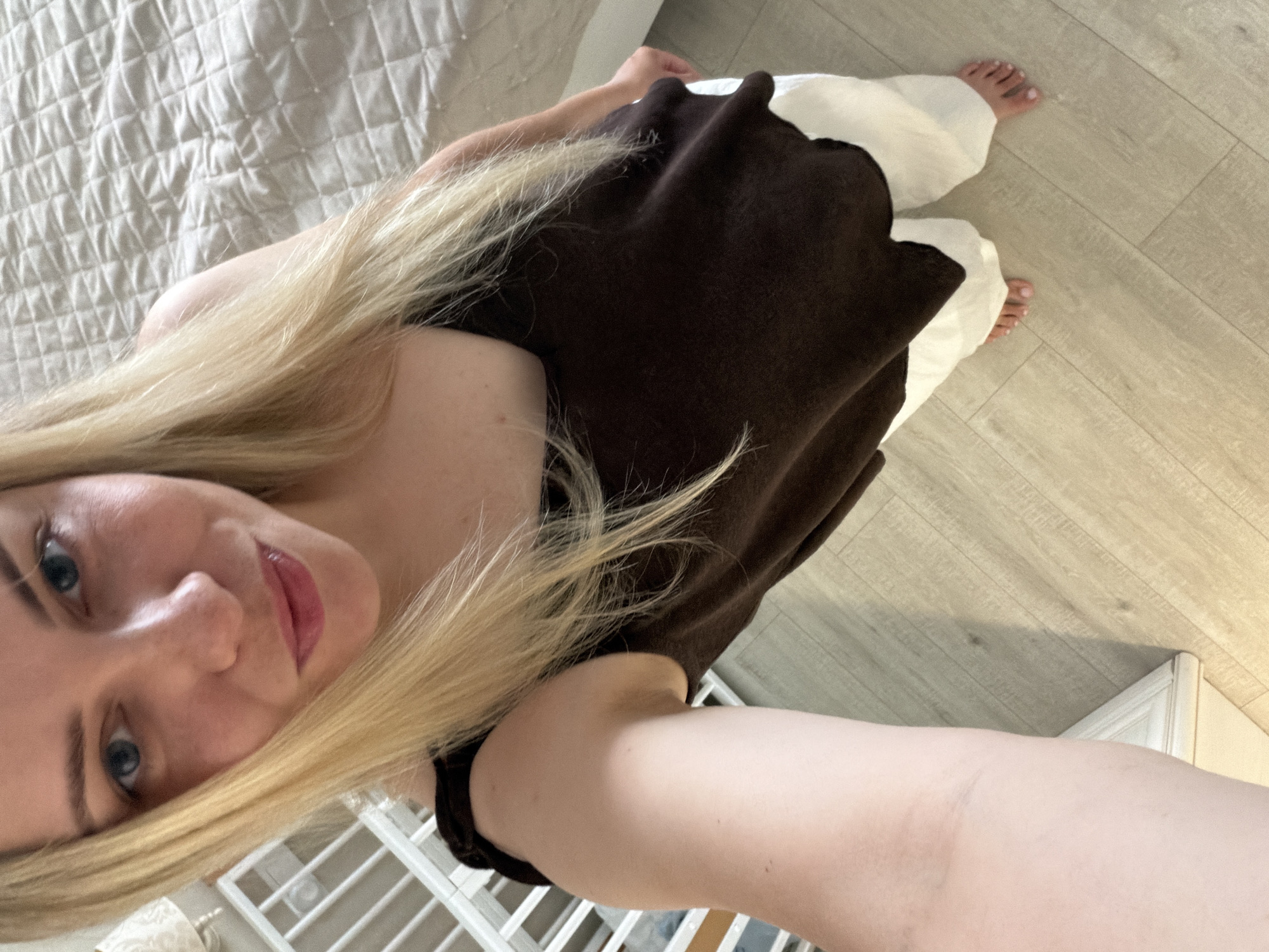 A young Caucasian woman with long blonde hair and blue eyes is taking a selfie in what appears to be a bedroom. She is wearing a brown tank top and white pants. She is standing on a light-colored wooden floor, and a bed with a quilted beige cover is visible behind her. To her left, a white crib or toddler bed is partially visible. The lighting suggests it is daytime, with natural light coming from an unseen source. The woman is looking directly at the camera with a slight smile. There are no other people or animals present in the image. The location is not specified as being in Votkinsk, Russia, or anywhere else.