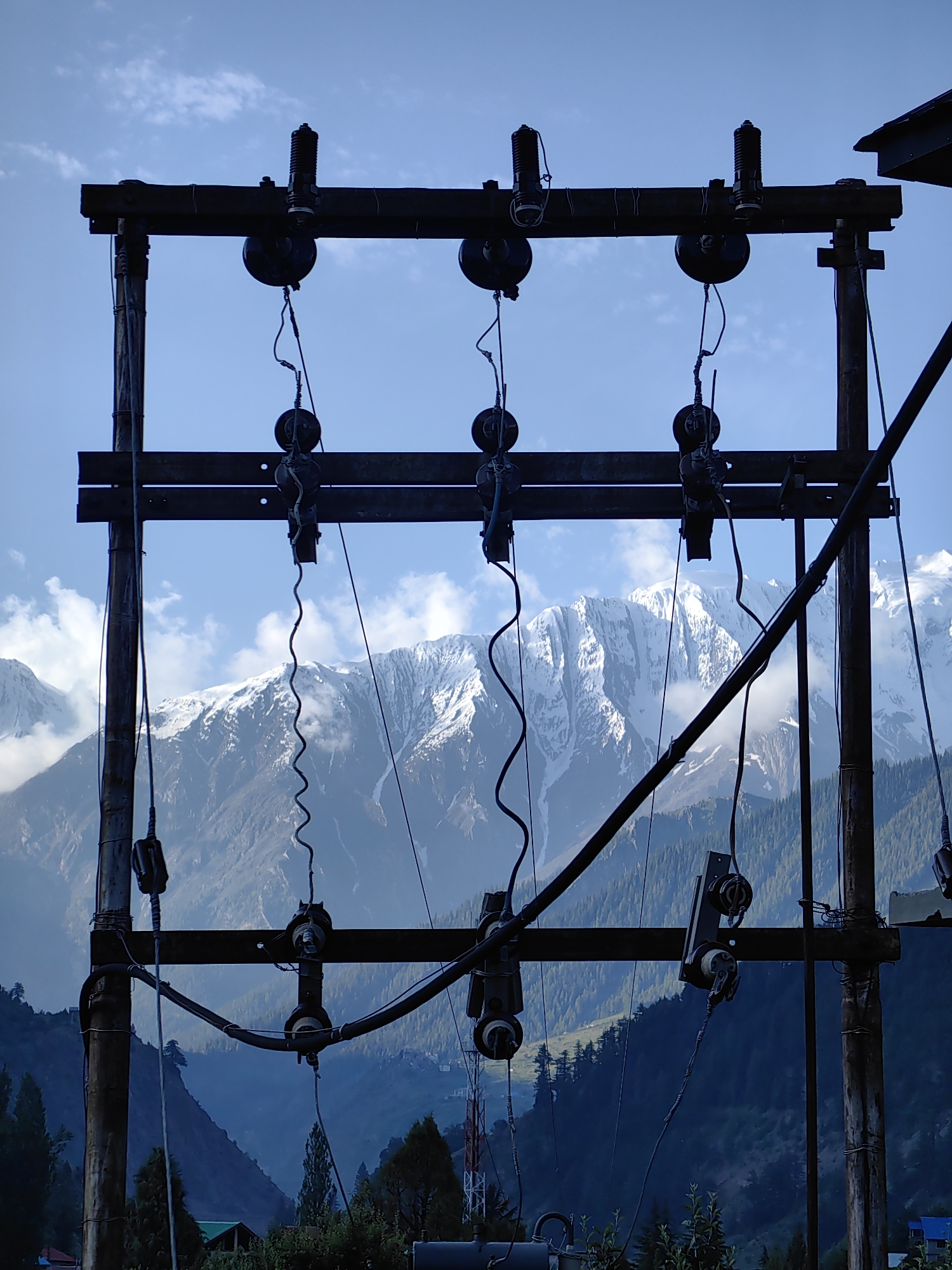 The image depicts a utility pole with electrical insulators and wires silhouetted against a backdrop of snow-capped mountains and a blue sky with scattered clouds. The pole structure is made of dark, weathered material, and several electrical components, including insulators and coiled wires, are visible. The mountains in the background are rugged and appear to be covered in snow, with forested slopes leading up to them. The overall scene suggests an outdoor environment, possibly in a mountainous region, where electrical infrastructure is present. There are no people or animals visible in the image, nor any actions or interactions taking place. The image has a high contrast between the dark foreground and the brighter background, with the sky and snow-covered mountains being the most illuminated elements. The location is not Udaipur, India.