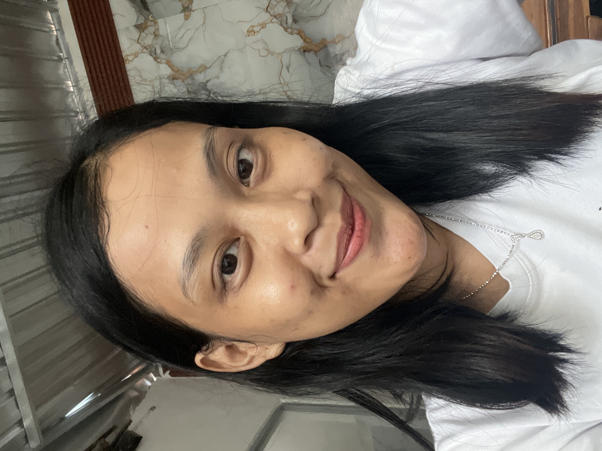 A woman is pictured in a close-up, frontal, selfie-style shot. She has dark, straight hair that falls past her shoulders and wears a white t-shirt with a delicate silver necklace featuring a teardrop-shaped pendant. Her face is turned slightly to her left, and she is smiling. The background appears to be indoors, with a corrugated metal ceiling visible in the upper left corner. To the right of the woman, a wall with a marbled pattern is discernible. The lighting suggests an indoor environment, possibly a home, and the overall scene is casual and personal. The exact location within Kecamatan Pati, Indonesia, is not specified by any visual cues in the image.