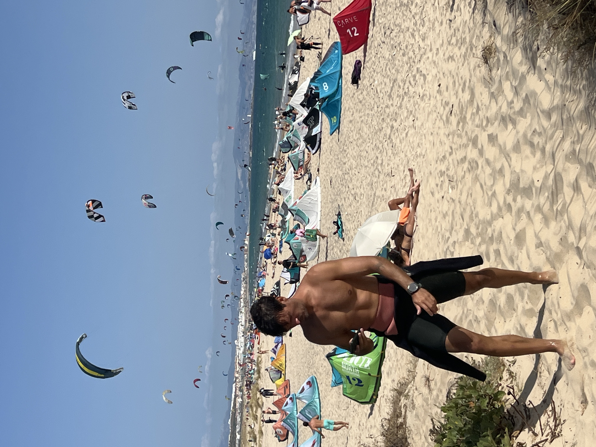 In Tarifa, Spain, a sandy beach is bustling with activity under a clear blue sky. The foreground features a shirtless man wearing black wetsuit shorts and a pink waistband, looking down at his phone. He has a watch on his left wrist. Behind him, the beach is populated with numerous colorful kitesurfing kites in various states of flight, with many people in the distance engaged in watersports and lounging on the sand. Several kitesurfing kites are set up on the beach, some with riders nearby. In the background, a town with white buildings lines the coast, with hills visible further inland. The ocean is a vibrant blue, with waves suggesting windy conditions suitable for kitesurfing.