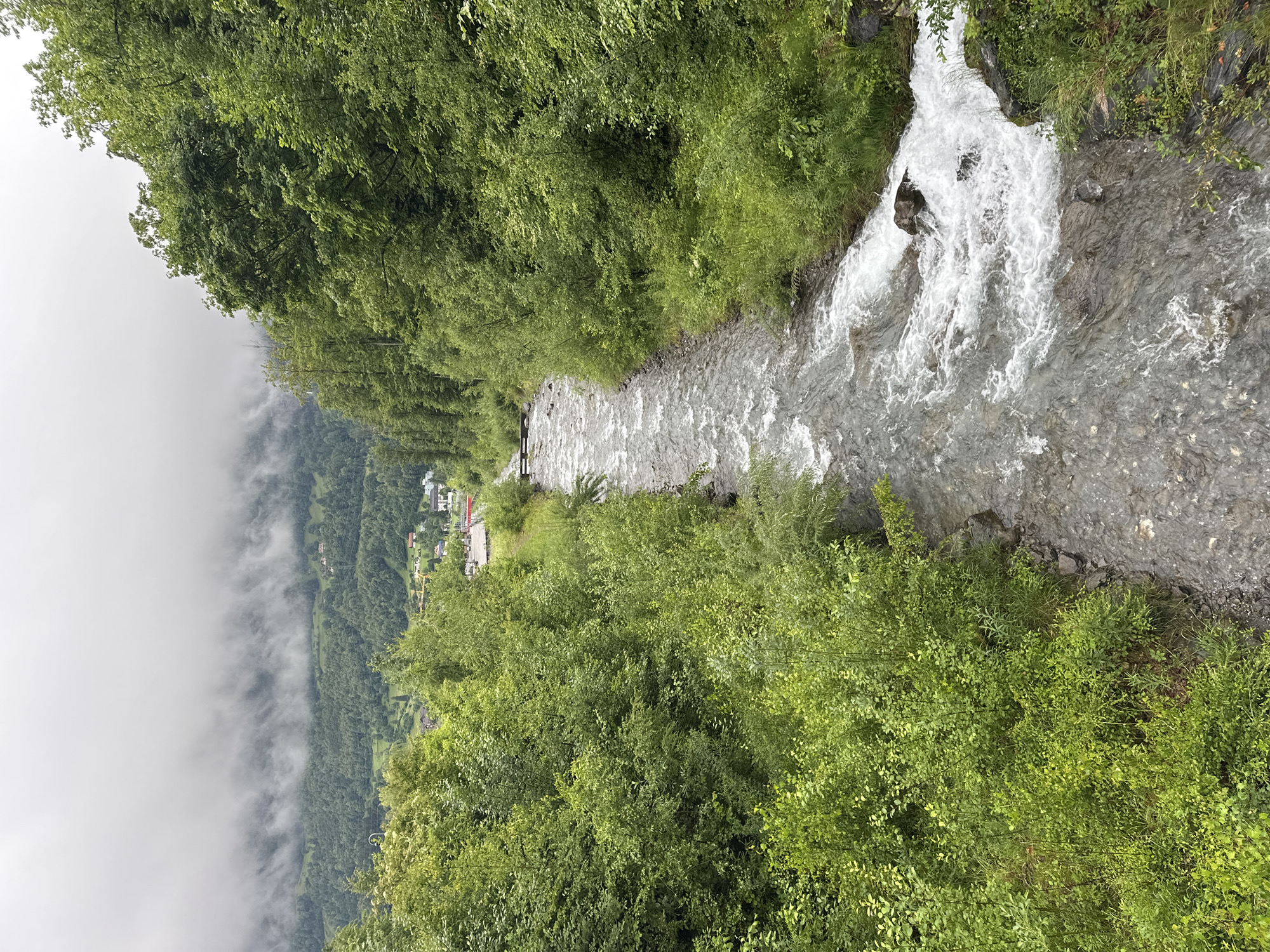 The image shows a fast-flowing river with rocky banks and lush green vegetation on either side. In the background, there are rolling hills covered in trees, with a village nestled in the valley. The sky is overcast, with mist or clouds obscuring the distant mountain peaks. The scene appears to be in a rural or mountainous area, possibly the Swiss Alps, given the mention of Hüsen, Switzerland. There are no people visible in the image. The river flows from the background towards the foreground, with whitewater rapids indicating its swift current.