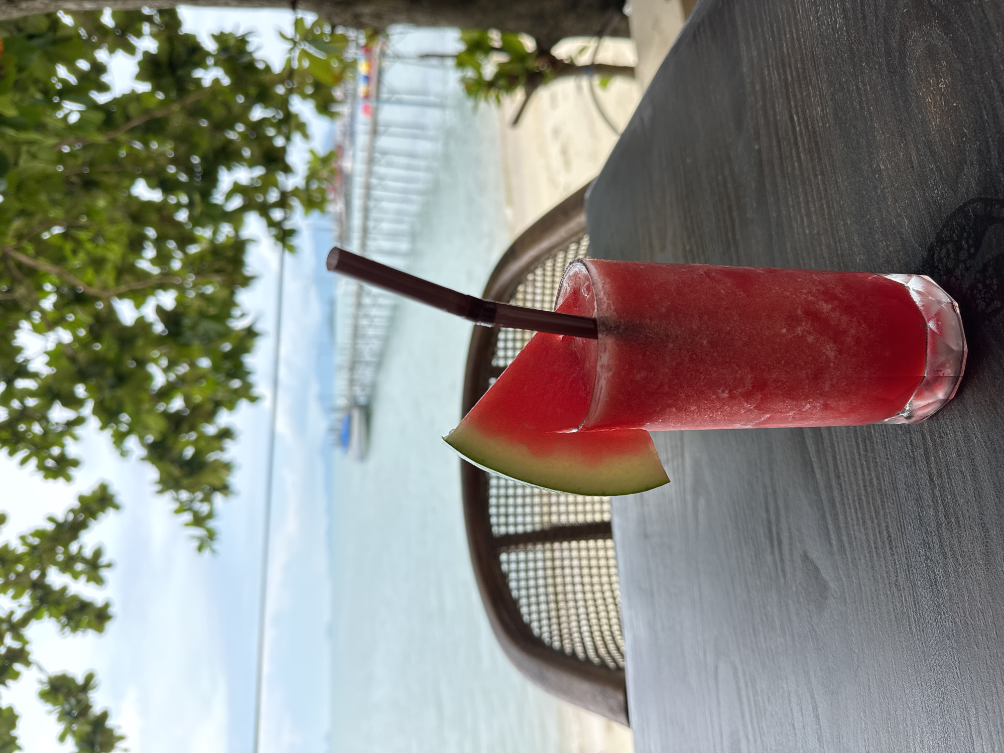 The image shows a watermelon smoothie on a grey wooden table. The smoothie is in a clear glass with a brown straw and a slice of watermelon on the rim. A woven chair is partially visible behind the glass. In the background, there is a beach with a long pier extending into the water and green foliage on the left. The scene appears to be set in Baan Bang Rak, Thailand, given the tropical beach environment.