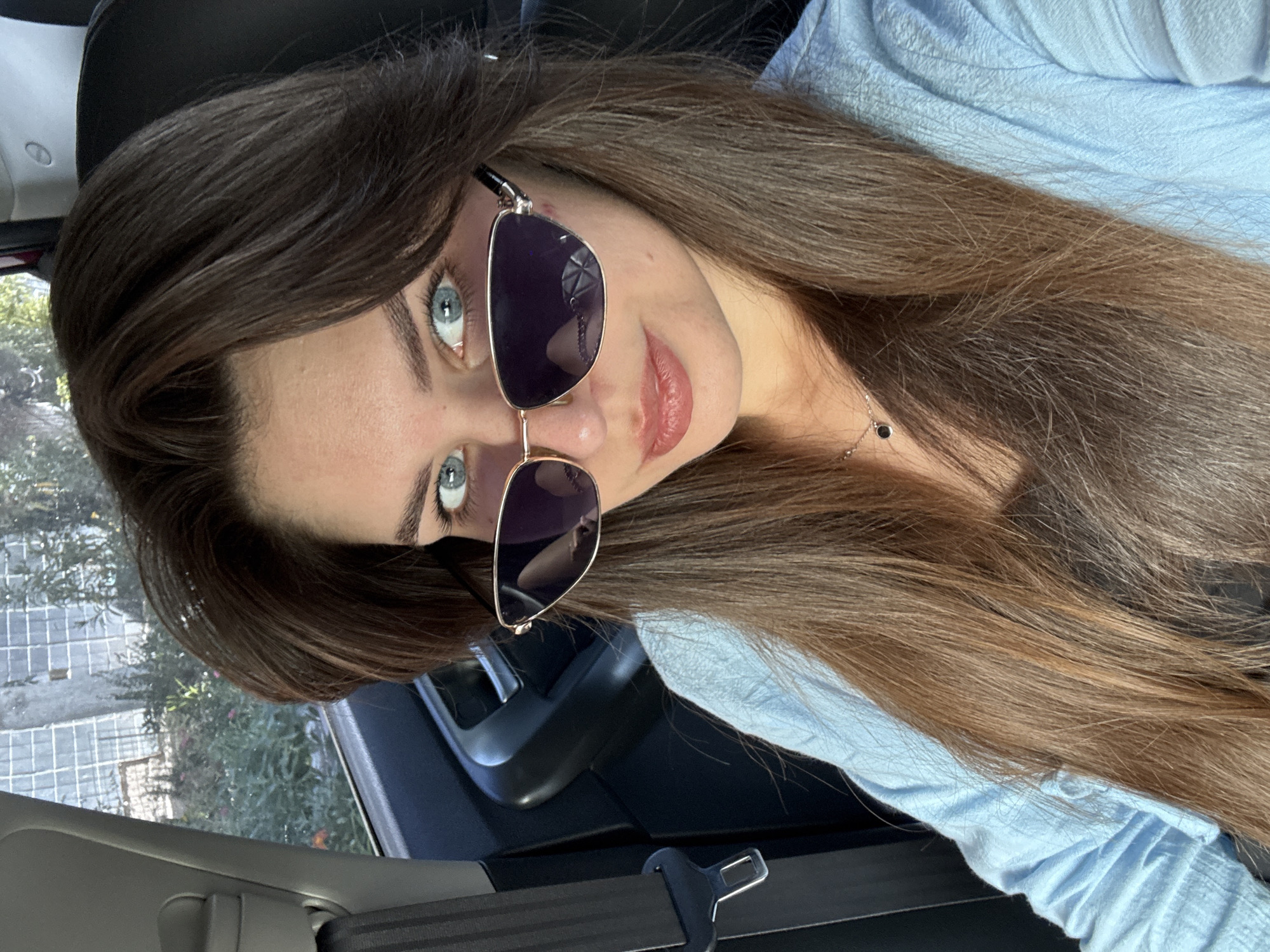 A woman with long brown hair and blue eyes is sitting in a car, looking at the camera. She is wearing sunglasses and a light blue shirt. The car appears to be in motion, with the scenery outside the window blurred. The seatbelt is visible on the left side of the frame.