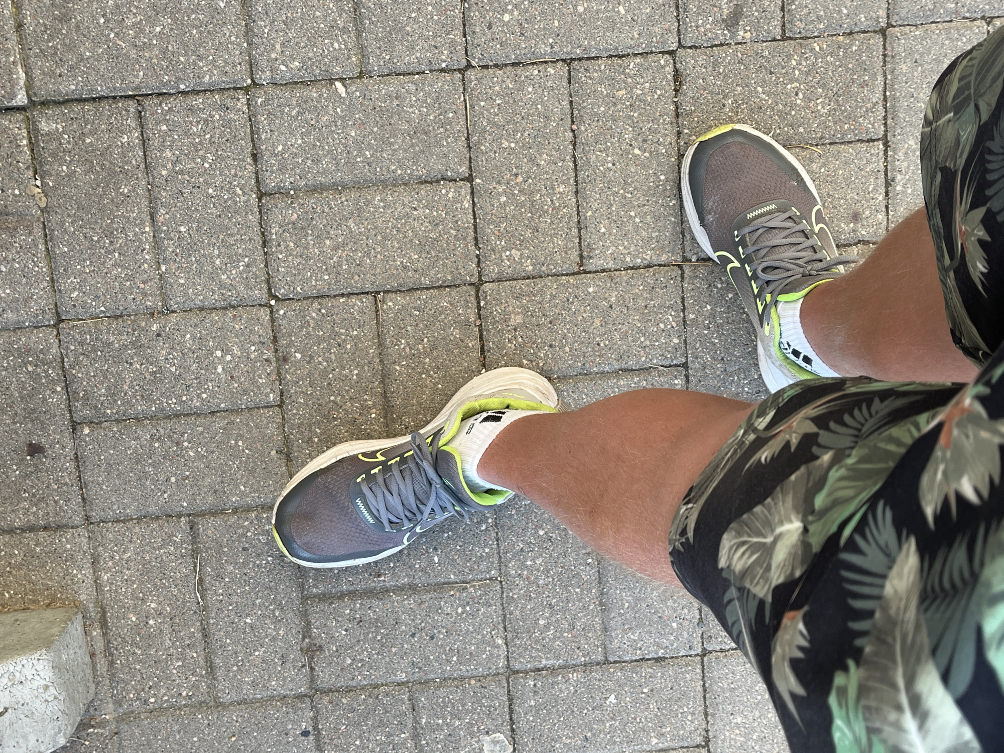 The image shows a person's legs and feet from a top-down perspective. The person is wearing dark shorts with a tropical leaf pattern and white athletic socks. They are also wearing gray running shoes with neon yellow accents. The person is standing on a sidewalk made of gray interlocking pavers. There is a rough concrete structure on the left side of the image. The scene appears to be outdoors, in a public space.