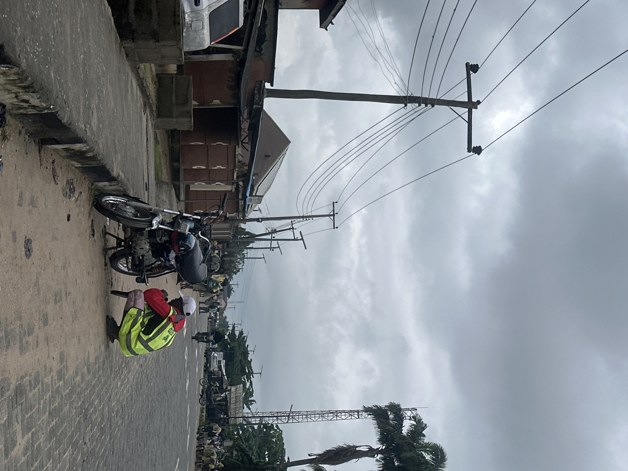 A person in a fluorescent yellow vest and white cap squats by a parked motorcycle on a paved street in Badagry, Nigeria. The sky is overcast. To the left of the street are buildings with reddish-brown walls and corrugated metal roofs. Utility poles with multiple wires line the street. In the background, other people and vehicles are visible.