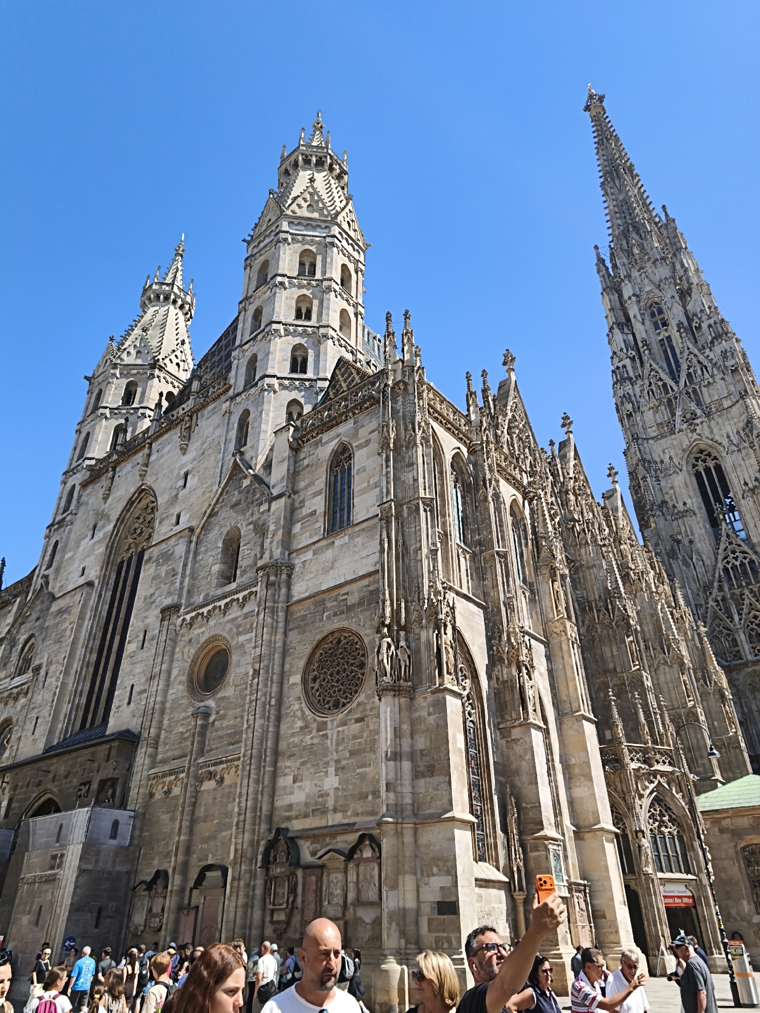 This is a photograph of St. Stephen's Cathedral in Vienna, Austria, taken from a low angle looking up at the facade. The cathedral is a Gothic structure made of stone, with intricate architectural details, including spires, towers, and arched windows. The sky is clear blue. A crowd of tourists is gathered in front of the cathedral, many of them taking pictures. In the foreground, a man is taking a selfie with his orange smartphone. The overall scene depicts a popular tourist attraction on a sunny day.