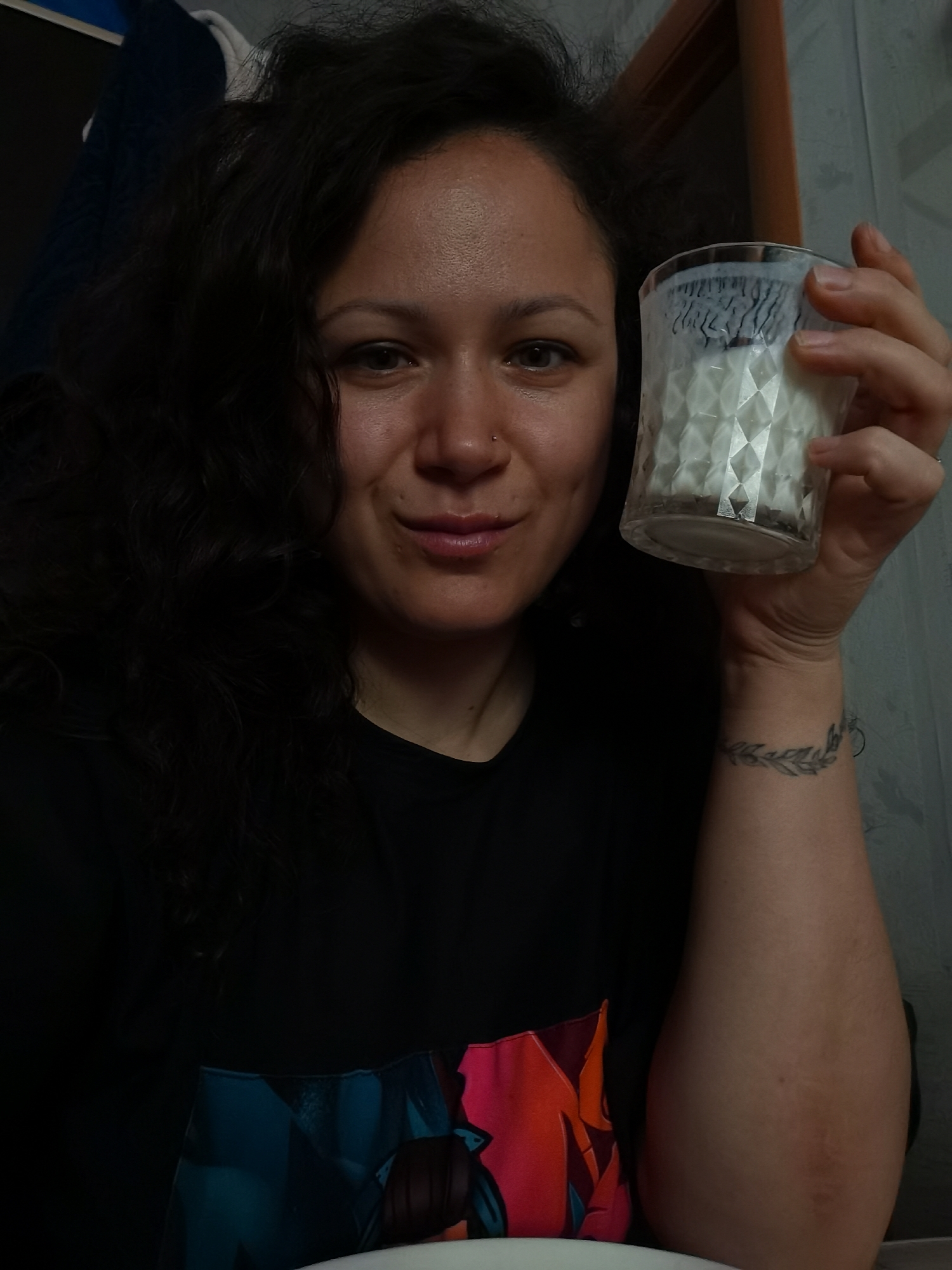 A close-up, indoor, selfie-style shot shows a woman with curly dark hair, holding a clear glass with a white textured interior. She is wearing a black t-shirt with a colorful abstract design on the lower portion of the frame. A tattoo of a leafy branch encircles her left wrist. The background is dimly lit, with a hint of a wooden frame on the right side and what appears to be a textured wall or curtain behind her. The woman's right hand is raised, holding the glass up towards her face, and she is looking directly at the camera with a slight smile. The image is undated and unlocated, and therefore it is not possible to specify the location in the city of Chaikovsky, Russia.