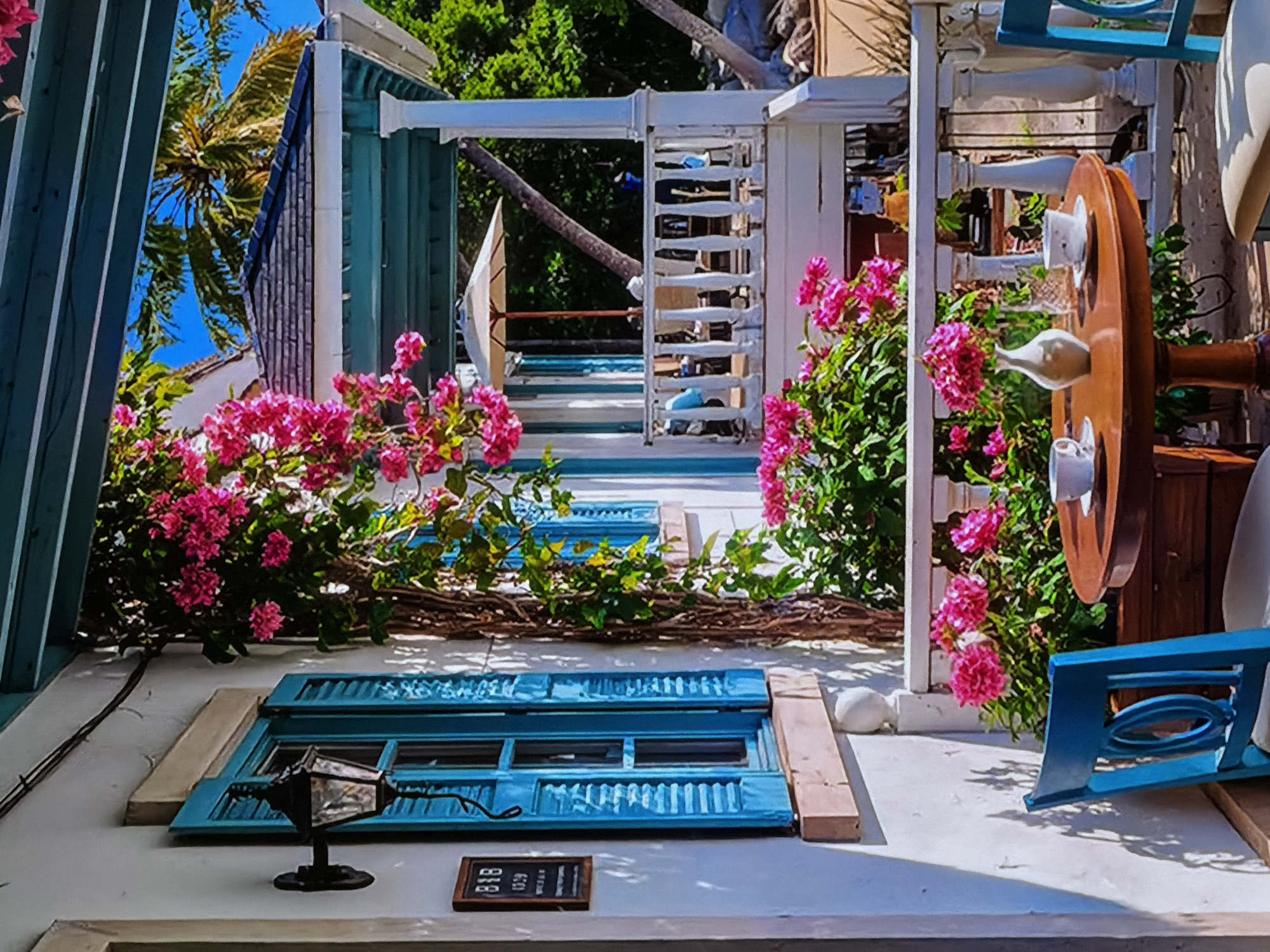 This vibrant image captures the colorful exterior of a building, likely a bed and breakfast or a themed establishment, located in Sofiivska Borshchahivka, Ukraine. The scene is bathed in bright daylight under a clear blue sky, with strong shadows indicating sunny weather.
The primary subjects include whitewashed walls contrasted with striking turquoise blue accents, such as functional window shutters and decorative architectural elements, possibly railings or pergolas. Abundant bushes of bright pink bougainvillea cascade throughout the scene, adding vivid splashes of color and a lush, overgrown aesthetic. Several palm trees are visible in the background, contributing to a tropical or Mediterranean atmosphere.
No people are present, depicting a tranquil and static moment. The focus is on the inviting and decorative nature of the architecture and landscaping.
Notable details include a blue-shuttered window on the lower part of the wall, below which a black, traditional lantern-style lamp is mounted. Above this window, intertwined bougainvillea and a natural woody branch create a charming decorative element. To the right, a large wooden wheel, resembling a ship's wheel, is mounted vertically and adorned with several small white ceramic bottles or vases. A distinctive blue chair-like structure with an intricate oval cutout design is positioned on the right side. On the ground, near the lower window, a small chalkboard sign is visible, displaying the text "B&B" and below it, what appears to be "1029" or "1229". A smooth, white, round object, possibly a stone or egg, rests on the ground nearby. The overall impression is one of cheerful decoration and a peaceful, inviting outdoor space.
