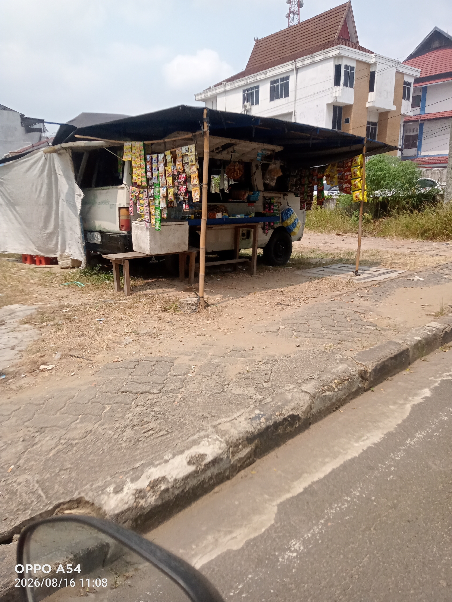 This is a photograph of a roadside kiosk in Jambi, Indonesia. The kiosk is set up on the side of a paved road, with a dirt and gravel area extending behind it. A white van serves as the base for the kiosk, with a dark tarp providing shade. Numerous small packages, likely snacks or cigarettes, are displayed hanging from the structure. A white cooler is placed on a wooden bench in front of the van. Behind the kiosk, there is a grassy area and a multi-story building with a distinctive red tiled roof. The sky is overcast. The timestamp on the image indicates it was taken on August 16, 2026.