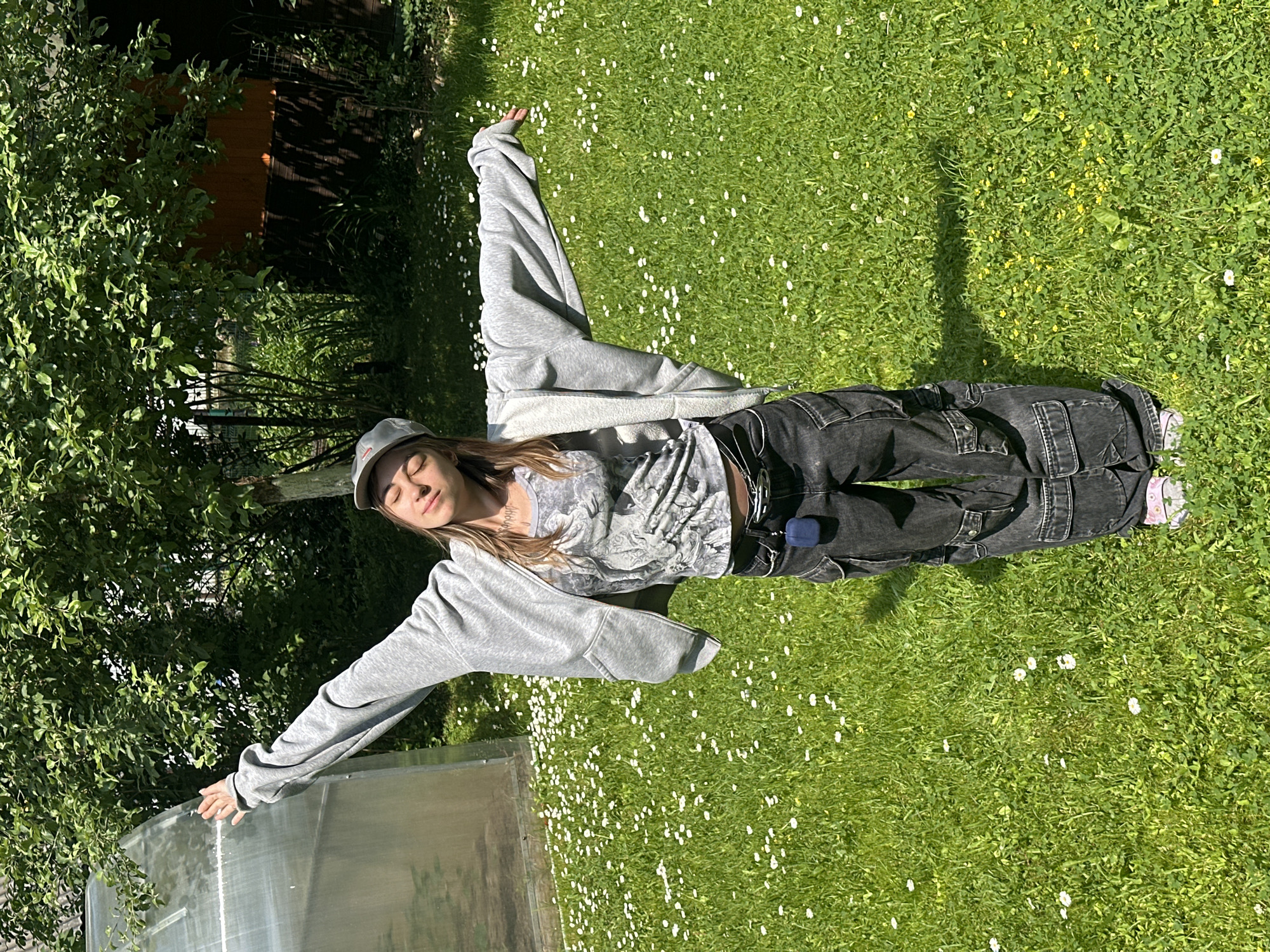 A young woman stands in a grassy yard with her arms outstretched in a sunny outdoor setting. She is wearing a grey hooded jacket over a graphic t-shirt, dark baggy cargo pants, a grey baseball cap, and pink slip-on shoes. A dark blue earbud case is clipped to her belt. The yard is covered in green grass and small white flowers, with a tree and a greenhouse visible in the background. The location is the city of Kozino, Russia.