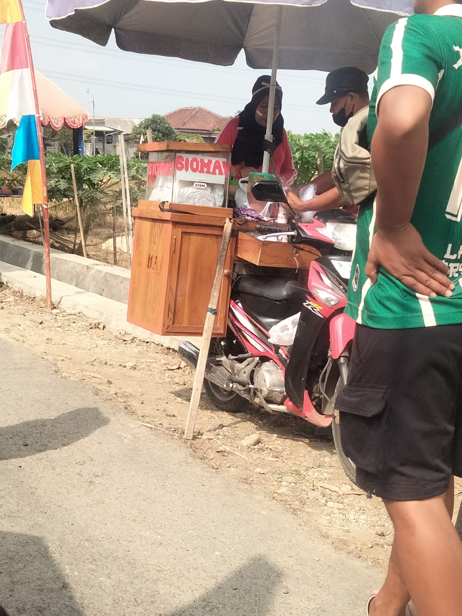 This image depicts a street scene in Ketanggung, Indonesia, where a mobile food vendor is operating. The vendor's stall is mounted on the back of a pink and black motorcycle. The stall consists of a wooden cabinet with a glass display case on top. The display case contains food items and is labeled "SIOMAY AYAM." Two individuals are visible near the stall. One, wearing a red shirt and headscarf, is attending to the food. The other, wearing a black cap and a dark face mask, is also near the stall, possibly a customer or an assistant. A third person, partially visible in the foreground, is wearing a green shirt and black shorts, their back to the camera. The setting is outdoors, with a paved road in the foreground and a dirt area with some vegetation and houses in the background. A large umbrella provides shade over the stall and the people. The overall impression is of a typical roadside food business in a rural or semi-urban Indonesian setting.
