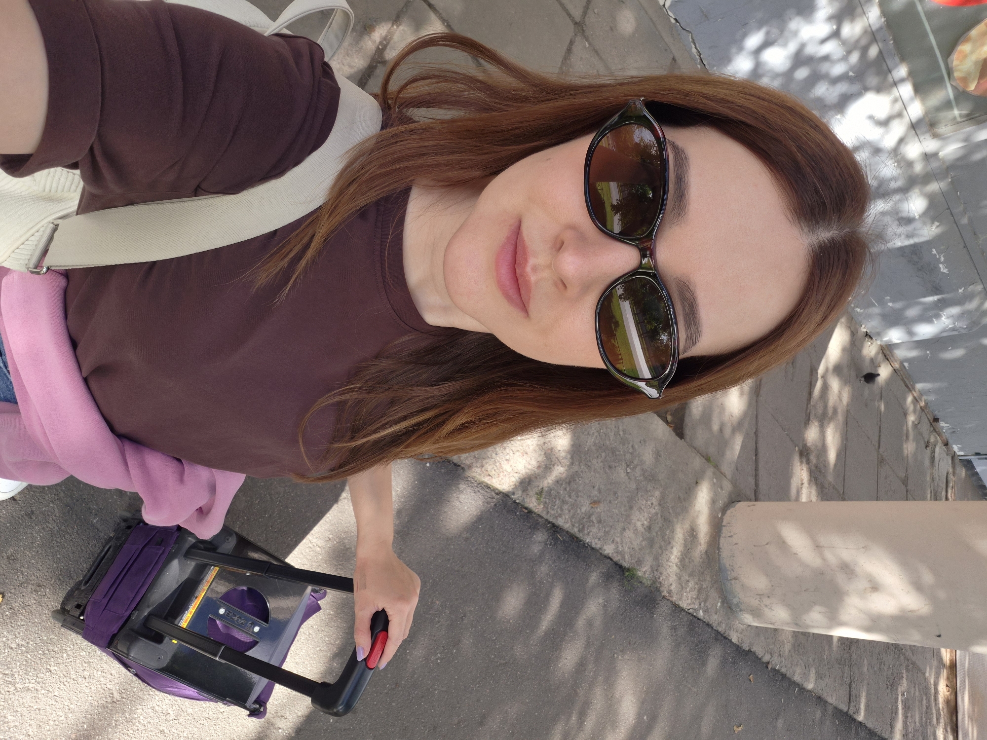 The image shows a woman taking a selfie outdoors in Saint Petersburg, Russia. She is wearing sunglasses and a brown t-shirt, with a pink sweater draped around her waist. She is holding the handle of a small purple rolling suitcase. The background features a concrete sidewalk, a light-colored pillar, and a gray wall with shadows from trees cast upon it. A pigeon is visible on the sidewalk in the distance.