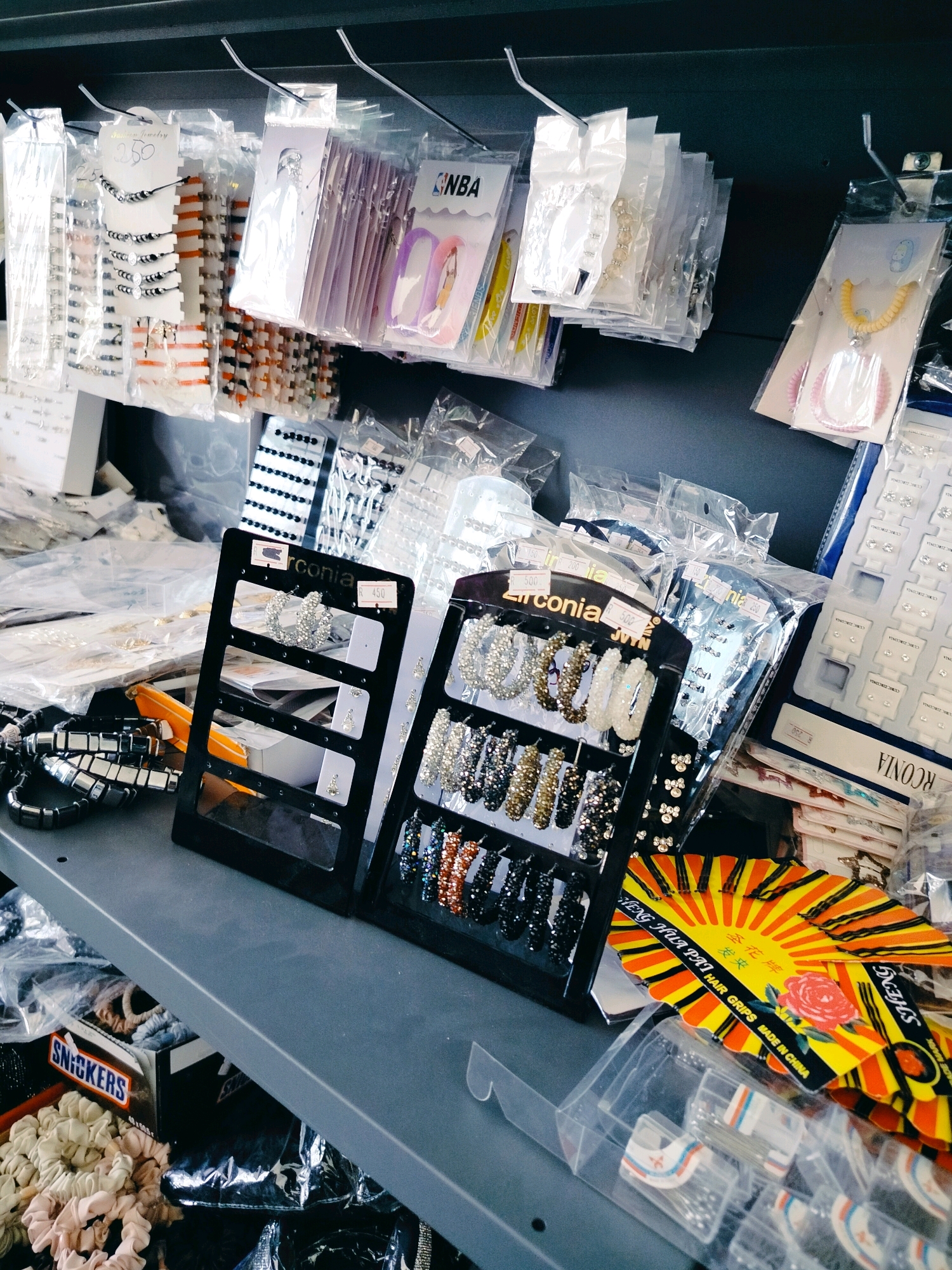 The image depicts a retail display of various accessories, likely for sale in a shop or market stall. The items are predominantly jewelry, including necklaces, bracelets, earrings, and hair accessories. They are displayed in clear plastic packaging, hung from hooks on a dark shelf or rack, and also arranged on black plastic display stands. Some of the jewelry is embellished with beads and rhinestones.
On the lower shelf, there are boxes of "Snickers" candy, which appear to be used as a container or prop. Scrunchies and other hair ties are also visible on this shelf, along with more packaged accessories. A vibrantly colored, fan-shaped display card with Chinese characters and an image of a rose is prominent in the foreground. The overall impression is of a busy and diverse collection of small, consumer goods. The image is taken from a slightly elevated, forward-facing perspective, looking down at the merchandise. The lighting suggests an indoor environment, possibly a market or small store. The specific location of Көлкент, Kazakhstan, is not visually evident in the provided image.