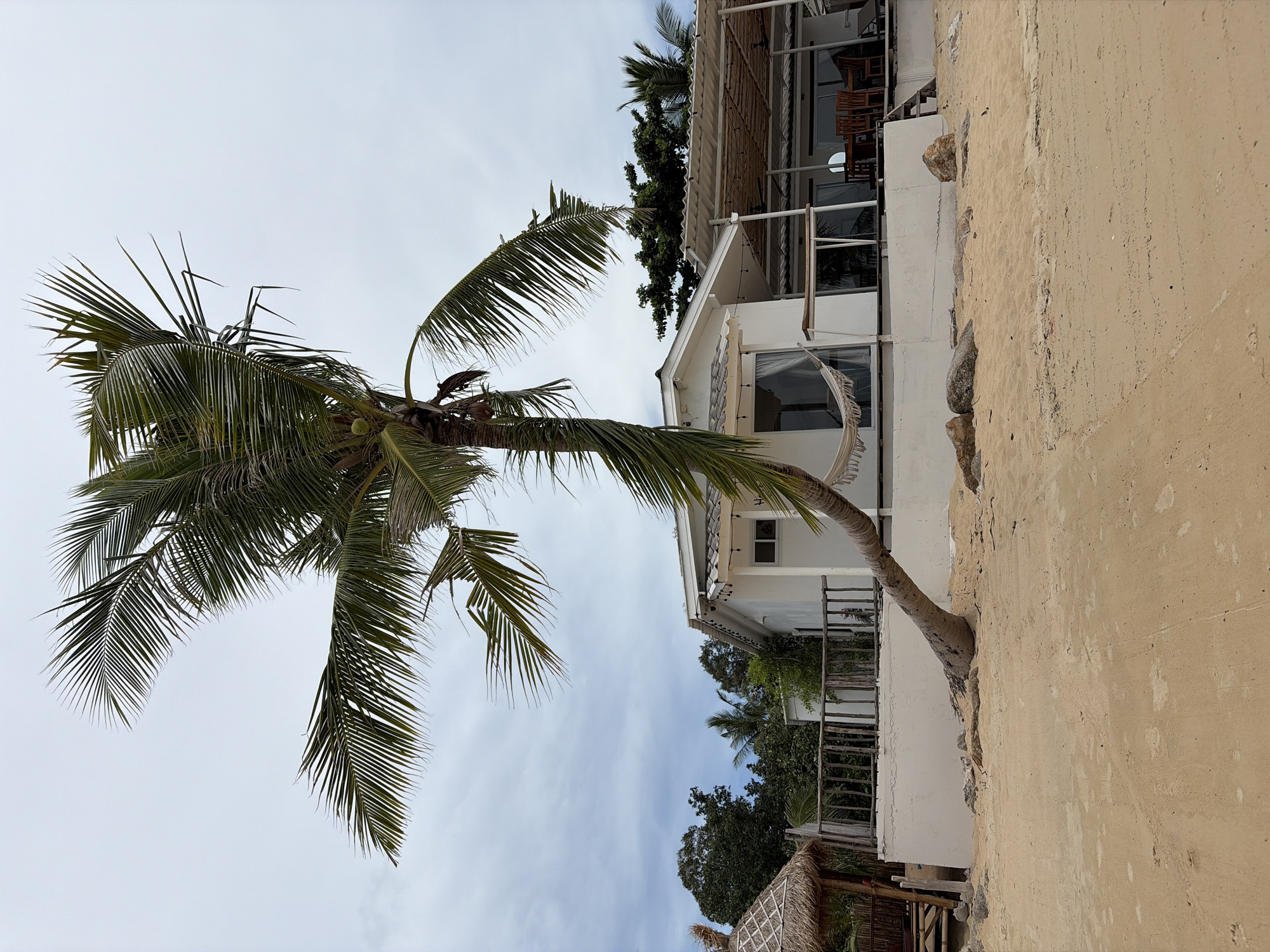 A full outdoor, eye-level shot depicts a sandy beach with a leaning palm tree in the foreground. The palm tree's trunk curves significantly, extending over the sand towards a white building. The building has a thatched roof hut to its left, a white wall surrounding its base, and a porch area with a covered roof. A hammock is visible hanging on the porch. The environment is overcast, with a sky filled with clouds. Lush green trees are seen in the background, behind the building. The image was taken in Baan Mae Nam, Thailand.