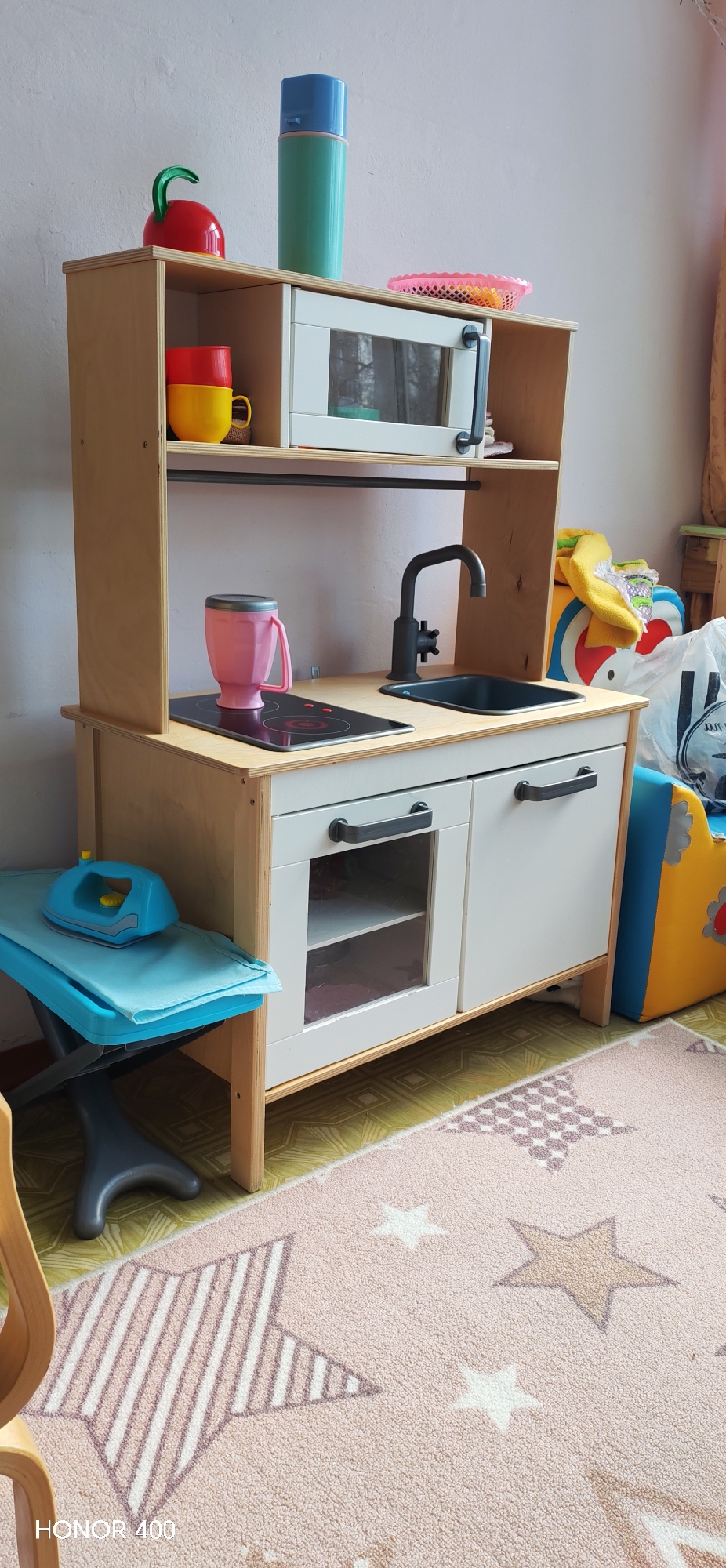 This indoor photograph captures a child's wooden play kitchen, situated in what appears to be a home in Noviy, Russia. The play kitchen features a light wooden frame with off-white cabinet doors and a built-in toy microwave with a transparent door and a black handle. The countertop includes a black toy sink with a matching faucet and a black stovetop with two red "burners."
Several colorful toy accessories are arranged on and around the kitchen unit. On the top shelf, a red kettle with a green handle and a tall blue and green thermos are placed. Below, a red cup is stacked on a yellow cup, and a pink woven basket sits near the microwave. A pink blender or tumbler is positioned on the stovetop.
To the left of the play kitchen, a blue toy ironing board with a light blue cloth and a blue toy iron rests on the floor. To the right, a colorful, soft children's item, possibly a chair or toy, in yellow, blue, and red, is partially visible, along with a white plastic bag. The floor is covered by a light pink rug decorated with white, pink, and brown stars, which lies over a greenish-yellow patterned floor covering. The ambient lighting suggests it is daytime. The text "HONOR 400" is visible in the bottom left corner. The scene depicts a well-equipped play area for imaginative activities.