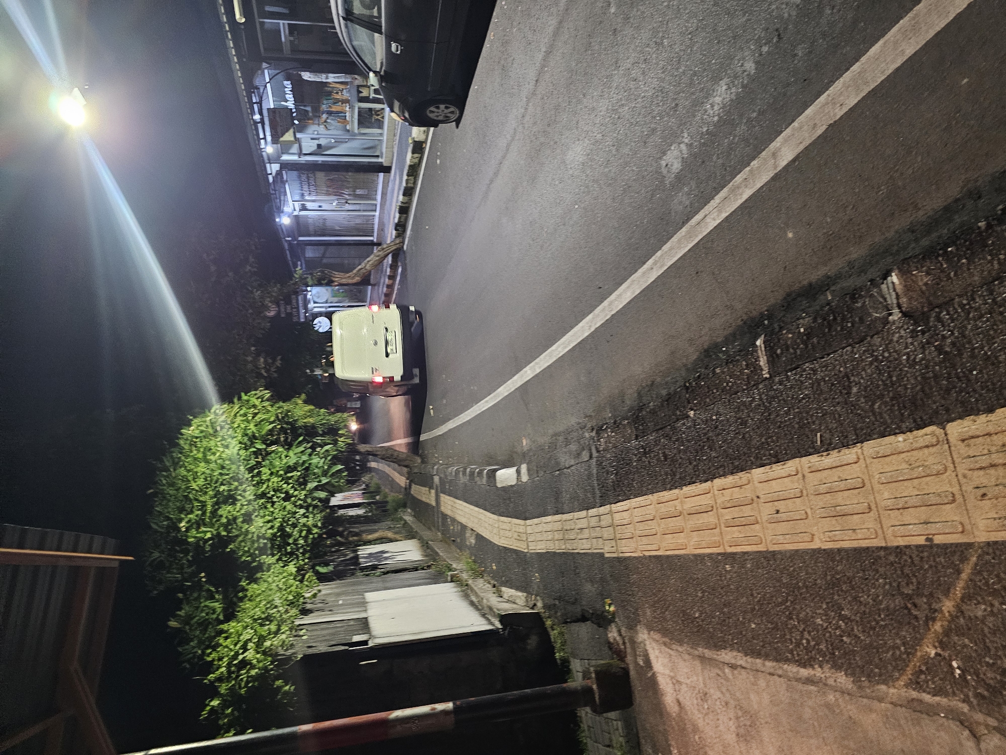 The image depicts a night scene on a street in Denpasar, Indonesia. A white line marks the edge of the road. A tactile paving section, with a textured surface of raised bars or domes, is visible on the edge of the road. Adjacent to the pavement is a row of buildings. On the other side of the road are green trees. A vehicle is partially visible at the top, and another is in the middle of the frame. A street lamp illuminates the scene from above. The image is rotated counter-clockwise.