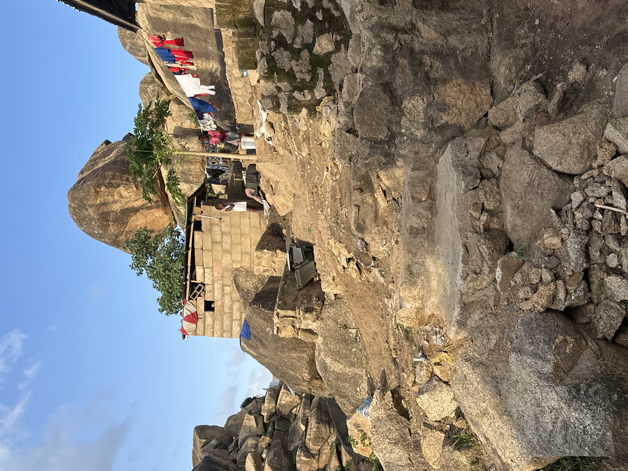 This photograph captures a vibrant scene of daily life set against a rugged, rocky landscape, characteristic of the Jos Plateau in Nigeria. Dominating the scene are massive, naturally weathered granite boulders, which serve as both the foundation and backdrop for human habitation. Integrated among these rocks are several rudimentary structures, primarily constructed from concrete blocks, indicating a community built directly into and around the natural rock formations.
Several individuals are visible, hinting at the human presence. One person in a white t-shirt stands near a block building, while further into the scene, figures in pink and blue garments are partially seen amidst their surroundings. The most prominent activity is the drying of laundry; a colorful array of clothes, including reds, blues, and whites, hangs on lines strung between rocks and structures, swaying gently under a clear sky.
The time of day appears to be late morning or early afternoon, with bright sunlight illuminating the scene and casting moderate shadows. The sky is a vivid blue with scattered white clouds, signifying fair weather. Sparse vegetation, including a tree with large green leaves (possibly papaya) and patches of grass and moss, thrives in pockets of soil. Household items such as buckets, patterned fabrics, and discarded materials are visible near the dwellings, underscoring the practical aspects of life in this environment. The scene conveys a sense of resourceful living, deeply intertwined with the unique geology of the region. No discernible text is present in the image.