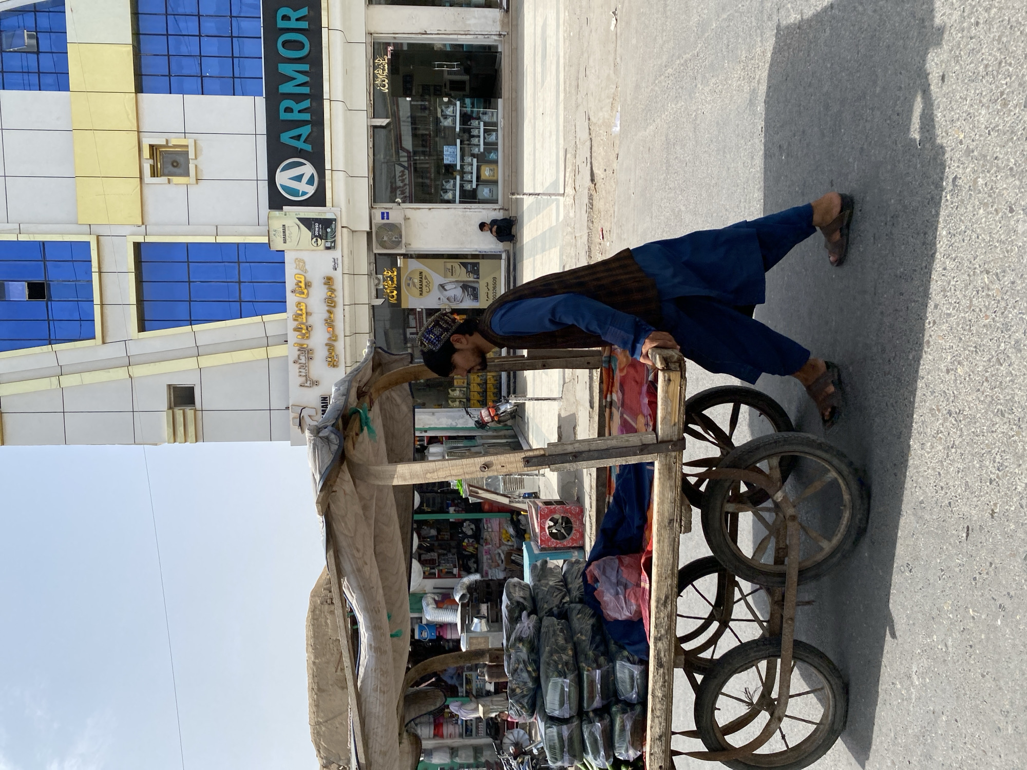 A man, wearing a blue shalwar kameez and a dark vest, pushes a wooden cart laden with goods. He has on a patterned cap and brown sandals. The cart is covered by a tarp and contains numerous wrapped items. The scene appears to be on a street in Kandahar, Afghanistan, with a modern building in the background. The building features blue glass windows, gold accents, and signage for "ARMOR." Another person, a child, sits on the sidewalk in front of the building. The street is paved and there is a shadow cast by the cart and the man. Other vehicles, including motorcycles, are visible in the distance.
