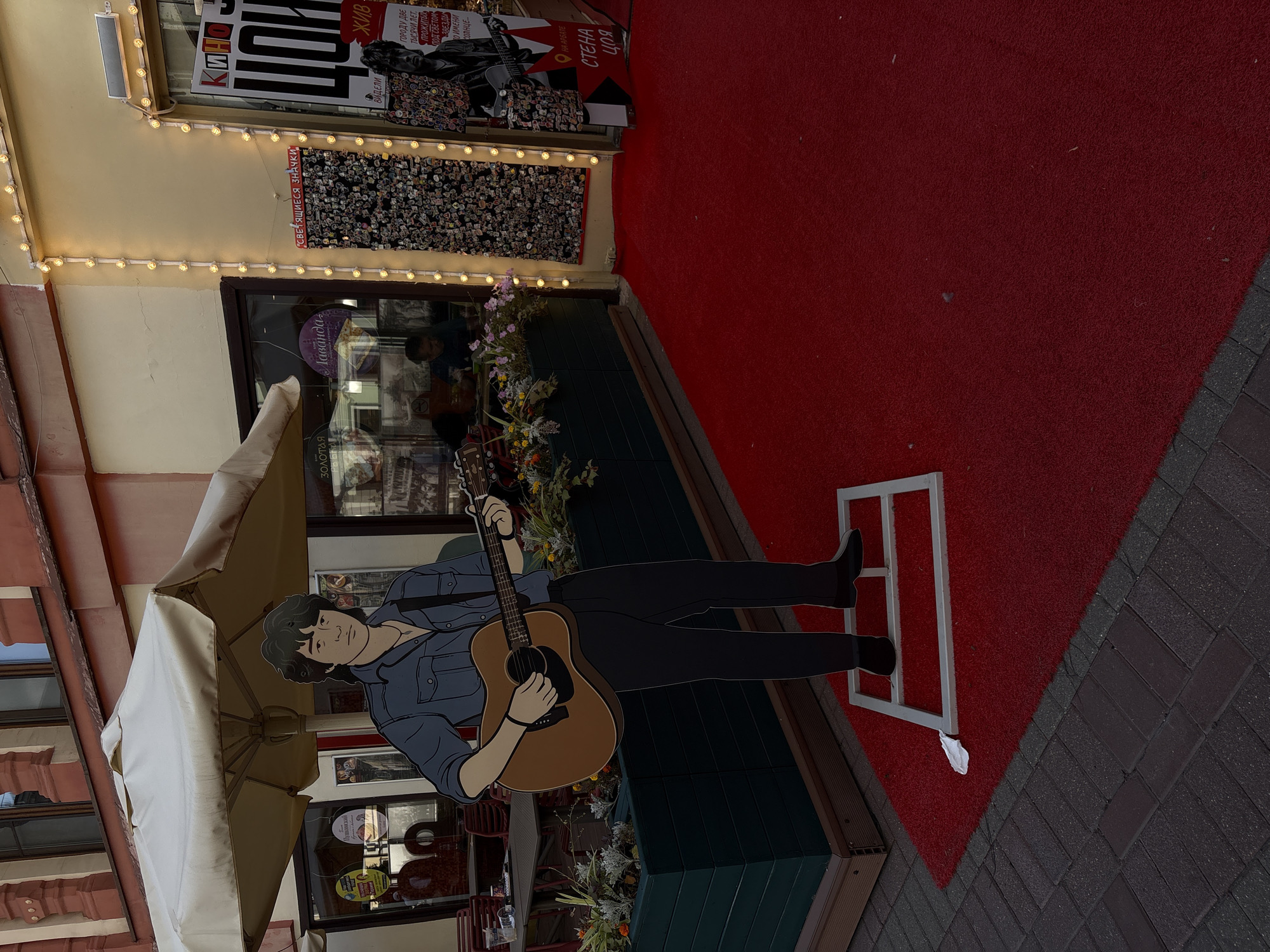 The image depicts a street scene in Moscow, Russia, likely in an area with shops and cafes. In the foreground, a red carpet extends across the cobblestone pavement, leading to the entrance of a building. A life-sized cardboard cutout of a young man playing an acoustic guitar stands on the red carpet. He is depicted with dark, wavy hair, wearing a blue denim shirt, black pants, and black shoes. He is strumming the guitar, which is brown. The cutout is supported by a white metal frame.
Behind the cutout, a building facade is visible. There is a large umbrella providing shade over an outdoor seating area with tables and chairs. The building features storefronts with windows displaying various items and posters. One poster, partially visible, has Cyrillic text and an image of a man playing a guitar. Another display features numerous small pins or badges attached to a dark surface, illuminated by string lights. Further details include planters with flowers, and a sign with Cyrillic text that reads "СТЕНА ЦОЯ", which translates to "Tsoi's Wall". This reference suggests a connection to Viktor Tsoi, a famous Soviet musician. The overall impression is a lively urban setting, possibly a pedestrian street or a cultural area.