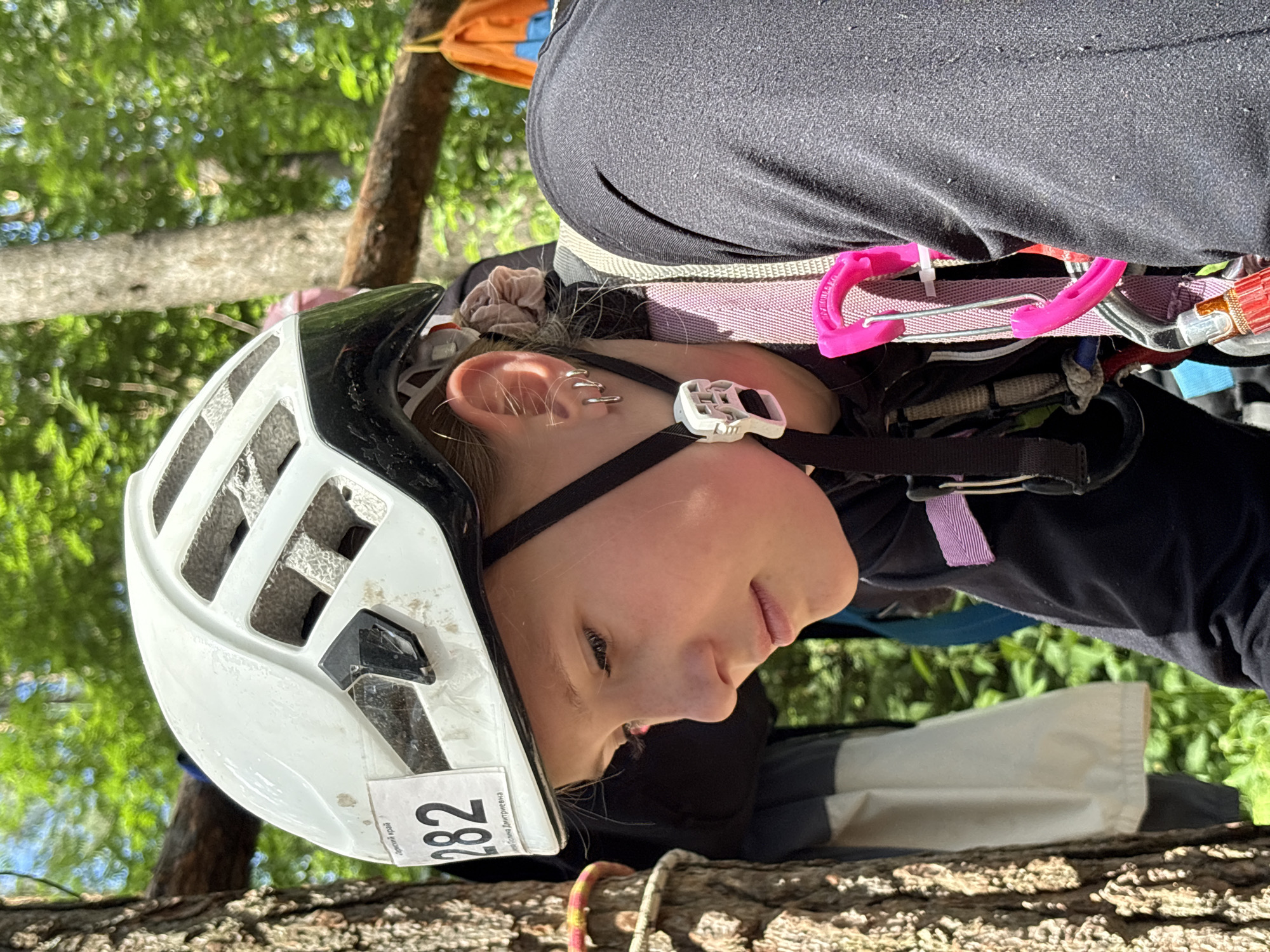 A young person wearing a white helmet with a sticker showing the number "282" is the central subject of this upward-facing, angled shot. They are looking down and to the right, with a focused expression, and their right ear, adorned with multiple piercings, is visible. The person is wearing a black top and a climbing harness, indicated by the visible straps, carabiners (one bright pink), and a metal buckle. The scene appears to be outdoors, with lush green foliage and trees in the background, suggesting a natural setting. The lighting indicates it is daytime, likely sunny, given the dappled light and shadows. The orientation of the image suggests the person might be climbing or suspended, possibly in a competition or activity that involves heights, such as tree climbing. The presence of the number "282" on the helmet could be an identification number for a participant in an event. The location is identified as Chaikovsky, Russia, which could imply a specific event or competition taking place there. The overall mood is one of concentration and engagement in an outdoor activity.