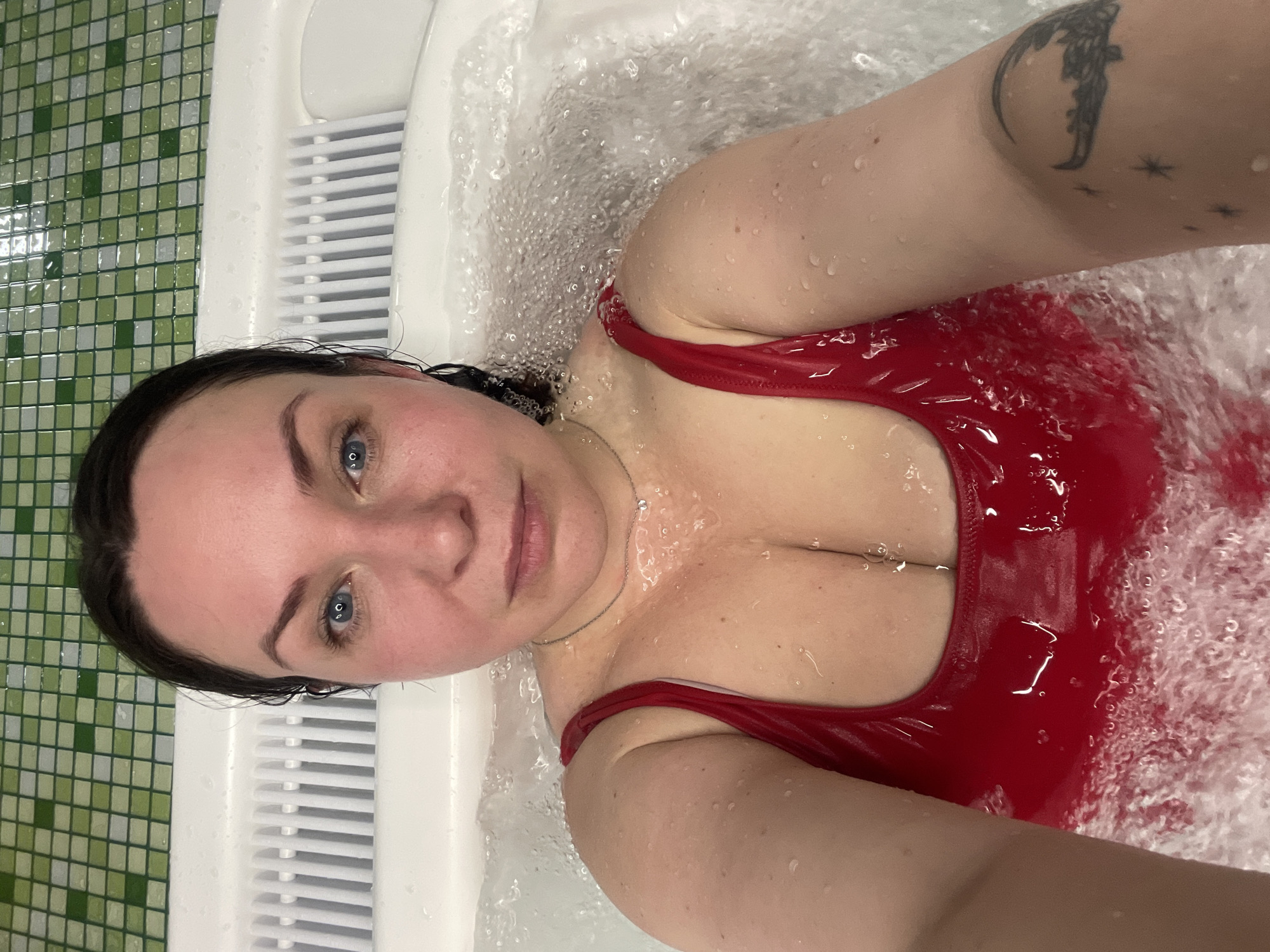 A woman with dark hair and blue eyes is shown in an indoor setting, possibly Kazan, Russia, wearing a shiny red swimsuit. She is immersed in bubbling water inside a white jacuzzi-style tub. A tattoo is visible on her upper left arm. The surrounding environment includes green and white tiled walls. The photograph appears to be a selfie taken from above.
