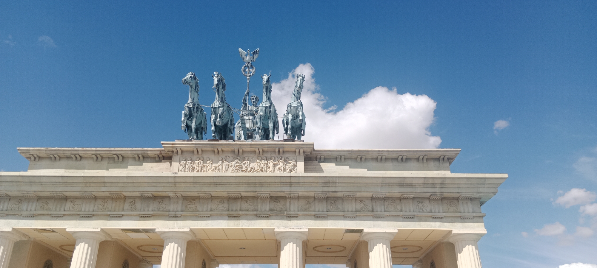 The image shows a close-up of the Brandenburg Gate in Berlin, Germany, not Torrejón De Ardoz, Spain. The gate is made of sandstone and is topped with the Quadriga, a statue of a chariot drawn by four horses and driven by Victoria, the Roman goddess of victory. The sky is bright blue with a few white clouds. The gate is a neoclassical monument that stands at the west end of the Unter den Linden, the boulevard that leads to the city palace. It is one of the most famous landmarks in Germany.