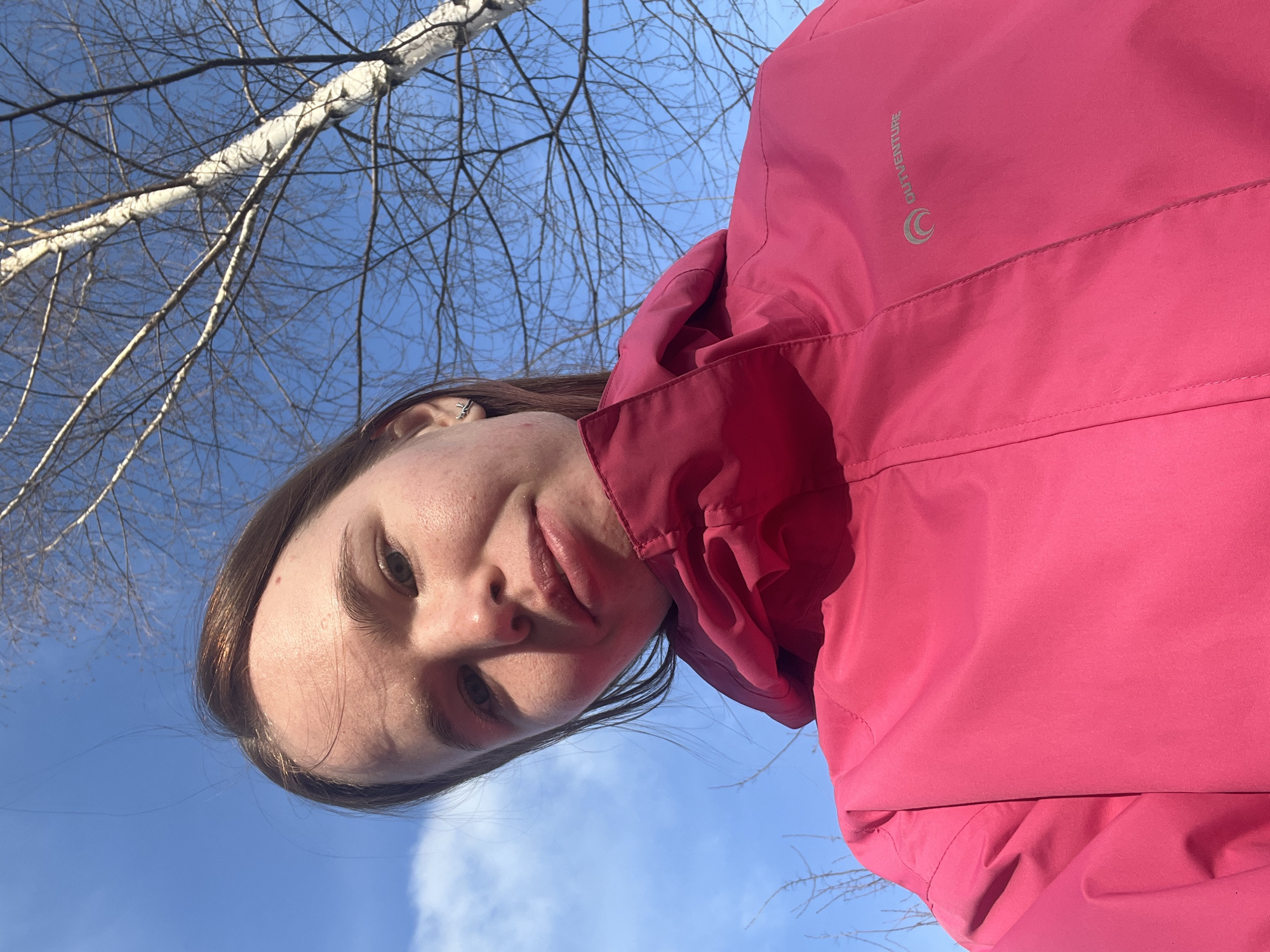 A person wearing a pink winter jacket takes a selfie outdoors. Bare tree branches and a blue sky are visible in the background. The location is Berezniki, Russia.
