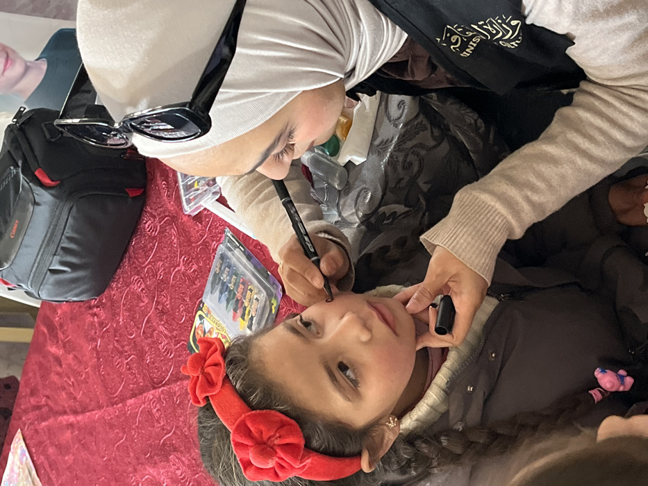 A woman wearing a hijab and black sunglasses draws on a young girl's face with a black marker. The girl wears a red headband with flowers. A black camera bag rests on a red tablecloth, along with crayons. The woman has a black apron with Arabic writing on it. She holds another black marker. The girl wears a gray jacket.