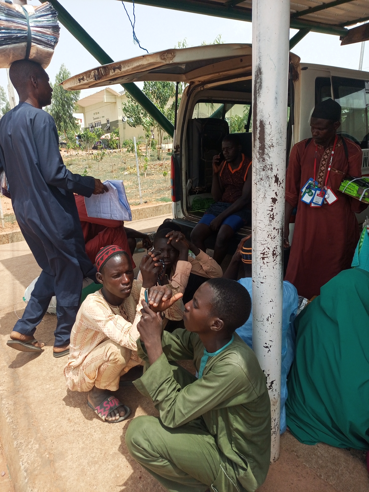 A group of people are congregated near a white van, likely in Gombe, Nigeria. One man in a dark blue robe carries a stack of items on his head and holds papers. A man in a brown robe with identification badges stands by the van. Inside the van, a person is on a cell phone. On the ground, several men are seated, with one holding a cell phone. The environment appears to be outdoors, possibly near a building labeled "RTEAN."