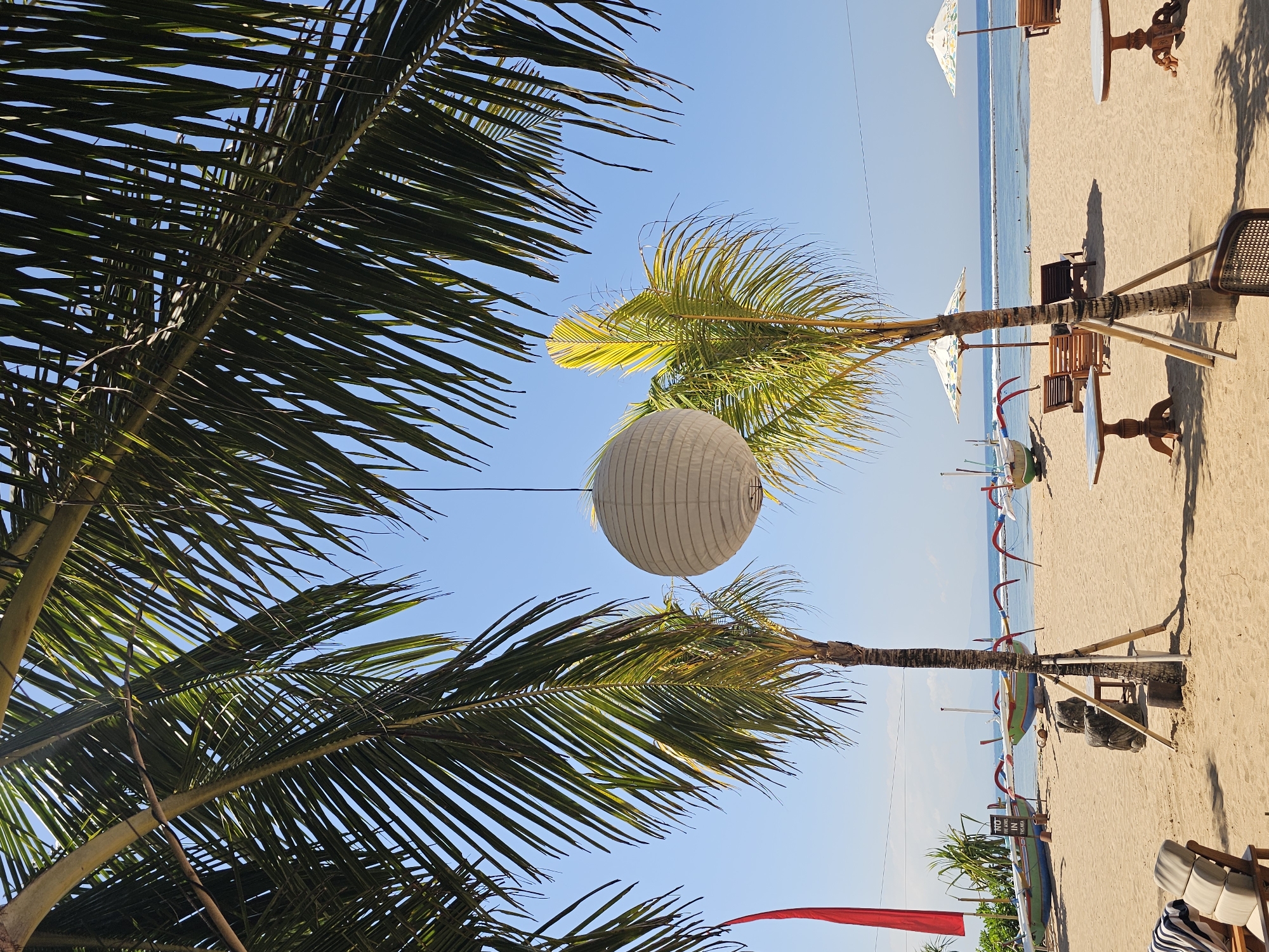 An outdoor, eye-level, medium shot captures a tropical beach scene in Denpasar, Indonesia, on a clear, sunny day. In the foreground, the sandy beach is dotted with lounge chairs and small tables, some with chairs. Palm fronds hang from the top of the frame, casting shadows on the sand. In the middle ground, several traditional Balinese jukung boats are pulled ashore, their colorful hulls contrasting with the beige sand. Two palm trees stand near the boats, supported by bamboo poles. A white, spherical lantern hangs suspended between the palm trees. In the background, the calm ocean stretches towards the horizon, with a few distant boats visible. A few people can be seen swimming or wading in the water. The overall atmosphere is serene and relaxed, typical of a beach resort.