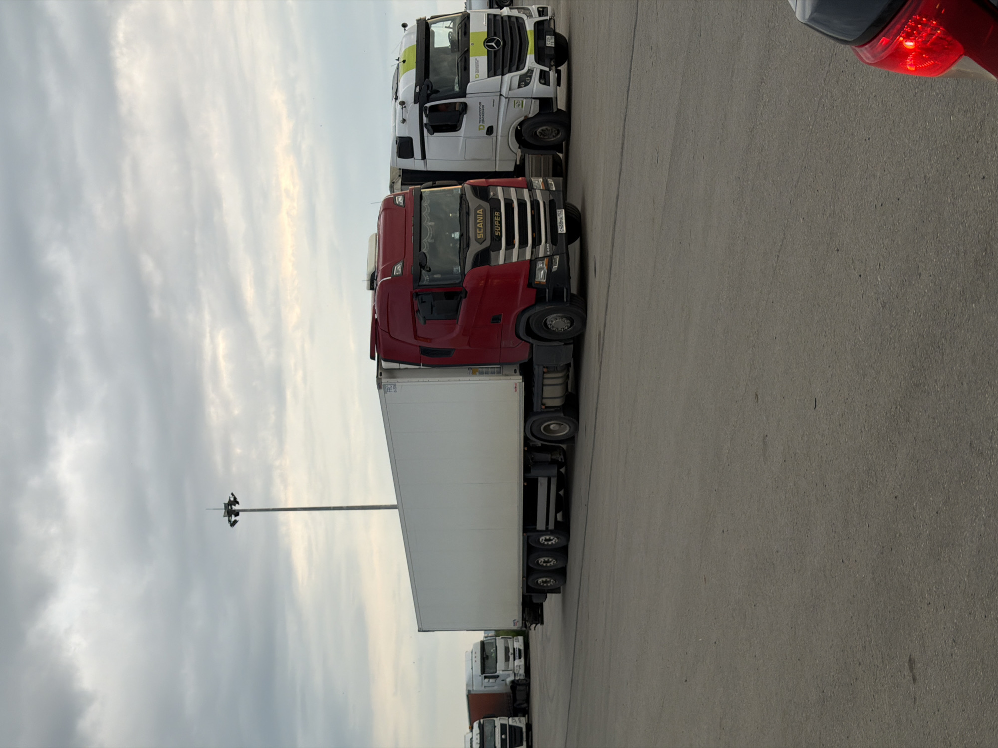 The image shows several parked semi-trailer trucks on a paved surface, possibly a parking lot or a loading area. The trucks are lined up side-by-side. The sky is cloudy, suggesting overcast weather, and the lighting indicates it is daytime, possibly late afternoon or early evening given the softness of the light.
The most prominent truck is a red Scania with a white trailer. Another truck visible is a white Mercedes-Benz with yellow and green accents on its cab, also pulling a white trailer. Several other trucks are partially visible behind these. The trucks appear to be stationary, suggesting they are parked, possibly waiting for loading or unloading.
In the background, a tall light pole with multiple lights is visible against the cloudy sky. The overall scene is utilitarian, characteristic of a logistics hub or industrial area. There is no visible indication of people in the image.
Visible text includes "SCANIA SUPER" on the grille of the red truck. The license plate on the red truck appears to be "C438MP 152". The white Mercedes-Benz has text on its side, with "ТЕХНОЛОГИЯ" and "ПРОДВИЖЕНИЯ" being partially legible.
The location is identified as Усть-Сыны, Russia.