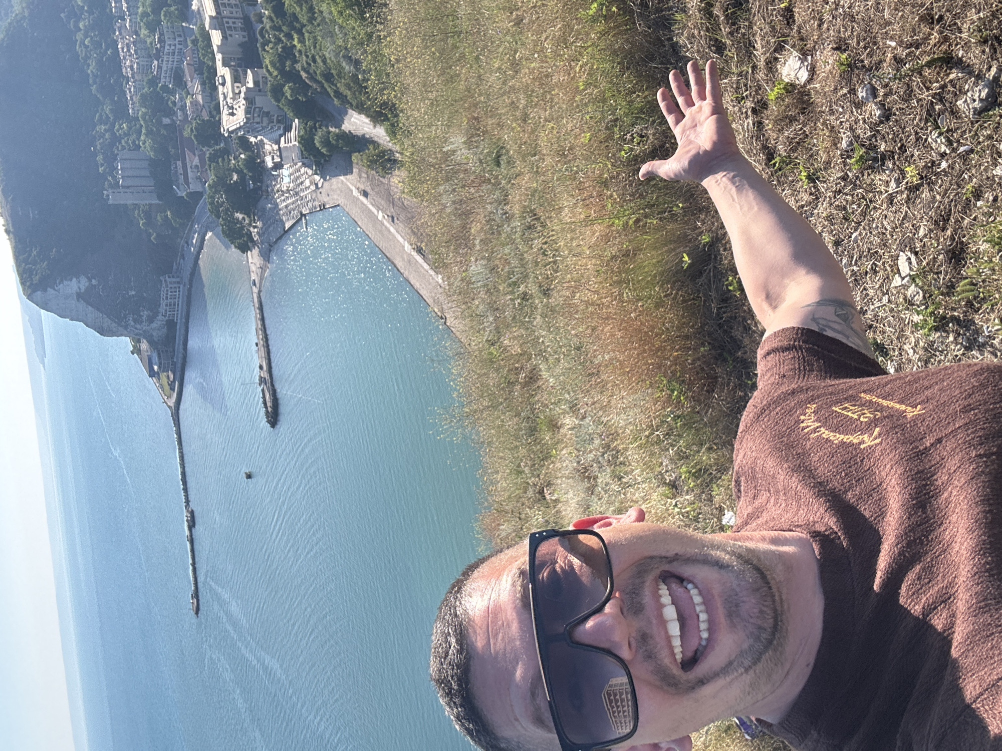 This is a portrait-oriented, slightly tilted photograph of a man taking a selfie. The man is wearing sunglasses and a brown, knitted sweater with text on the right chest that reads "Tropical Vibes 1977". He has a wide smile, showing his teeth, and appears to be outdoors. His left arm is raised, with his hand open, in a wave or greeting.
The background shows a dramatic coastal scene. To the left, there is a body of turquoise water, likely the sea, with a long pier extending into it. Along the coast, there are white cliffs and buildings nestled amongst lush green trees, suggesting a town or city located on the hillside. The terrain in front of the man is covered in dry grass and sparse vegetation, with some scattered rocks.
The sunlight suggests it is daytime, likely a bright and clear day, given the strong shadows and the sparkle on the water. The overall impression is of a cheerful and adventurous individual enjoying a scenic view. The location cues, including the cliffs and the style of buildings, are consistent with coastal towns in regions like Kavarna, Bulgaria.