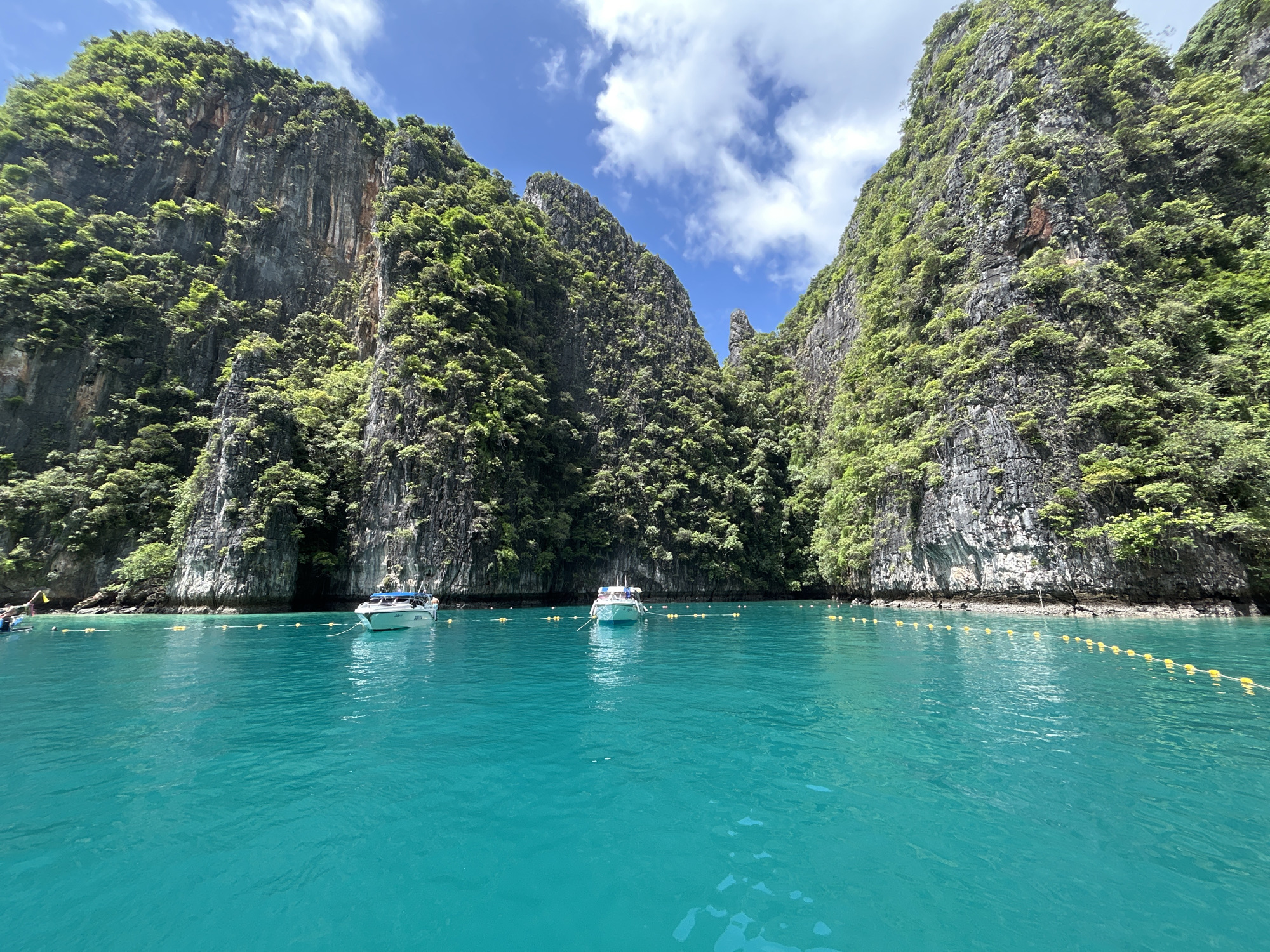 In Ao Nang, Thailand, a serene seascape unfolds, dominated by towering limestone cliffs cloaked in lush green foliage. The cliffs rise dramatically from the crystal-clear, turquoise waters of a bay, with their rugged surfaces etched by time and nature. The water is calm, with gentle ripples reflecting the bright blue sky, which is dotted with fluffy white clouds.
Several boats are anchored in the bay. Two speedboats are visible in the mid-ground, their white hulls contrasting with the vibrant water. One of these boats is positioned closer to the shore and is secured by a line of yellow buoys that mark a boundary in the water. Another smaller boat is further out, and a few individuals can be seen on it. The overall impression is one of tranquility and natural beauty, characteristic of the stunning coastal landscapes of Ao Nang.