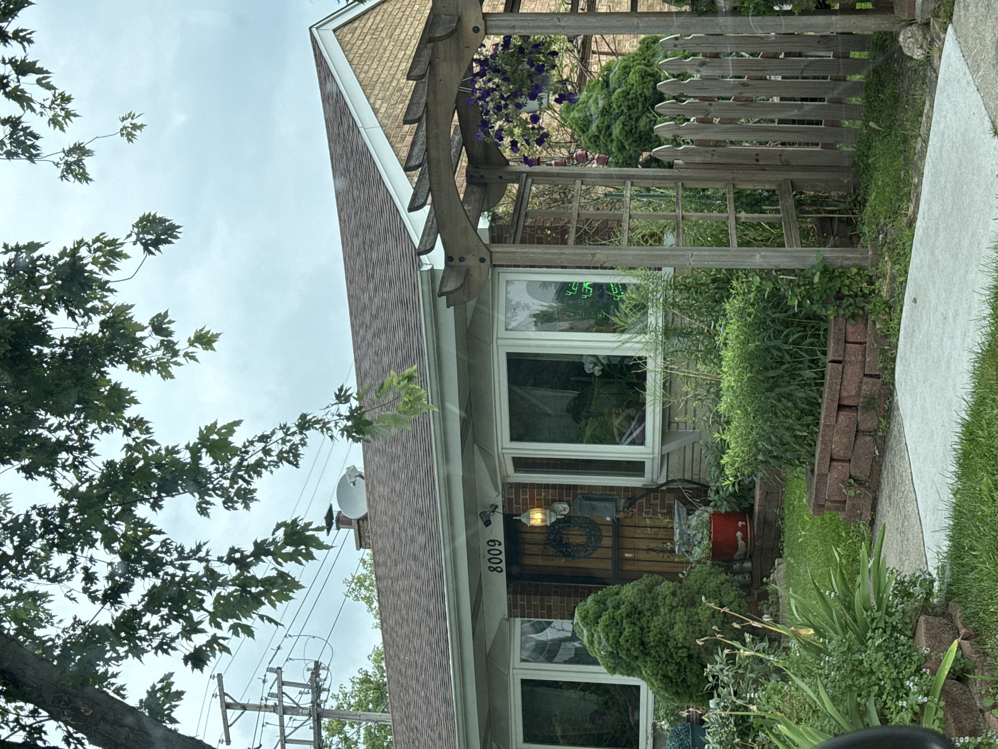 The image shows the front of a residential house located at 8009 in Skokie, Illinois, United States of America. The house has a brick facade, a brown shingled roof, and bay windows. In front of the house, there is a garden with various plants, shrubs, and flowers. A wooden pergola with hanging plants stands to the right of the entrance, and a picket fence encloses a portion of the yard. The sky is overcast. A tree with green leaves partially covers the top of the image, and utility poles and power lines are visible in the background. There are no people present in the image.