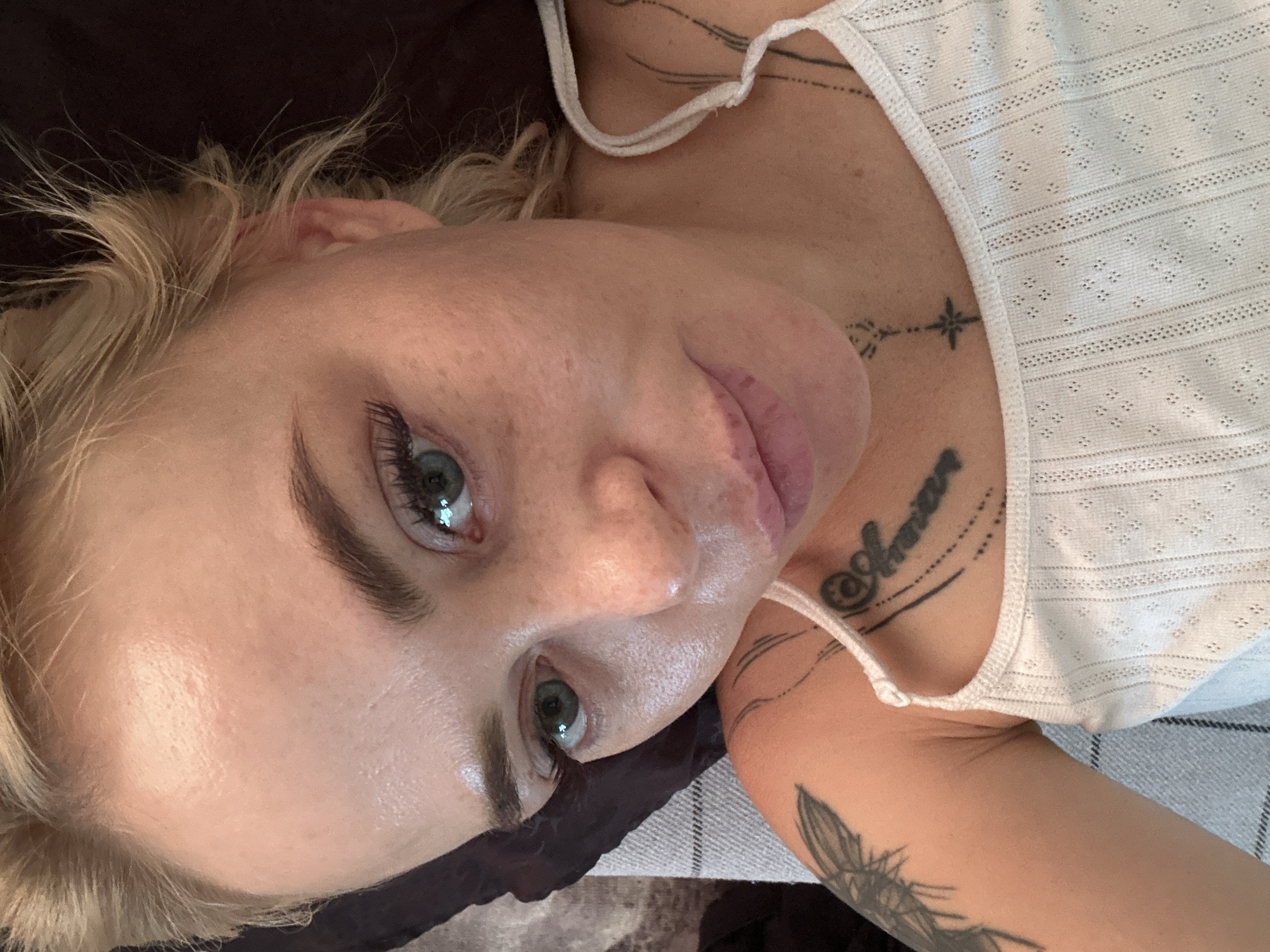 The image is a close-up selfie of a young adult, likely female, with blonde hair and fair skin. She is looking directly at the camera with a neutral expression. Her eyes are green and she has noticeable eyelashes. She is wearing a white, sleeveless tank top with a textured pattern. There are tattoos visible on her left shoulder and chest. The background is out of focus, suggesting an indoor setting. There are no visible clues to indicate a specific location within a city.
