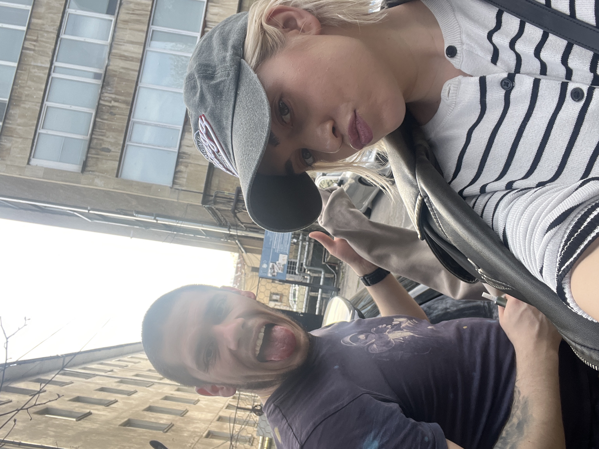 Two people pose for a selfie. A woman wearing a gray cap, striped cardigan, and black leather jacket is in the foreground, while a man with a purple T-shirt is behind. The man is sticking his tongue out. The location is Sofia, Bulgaria, as evidenced by the architectural style of the building in the background.