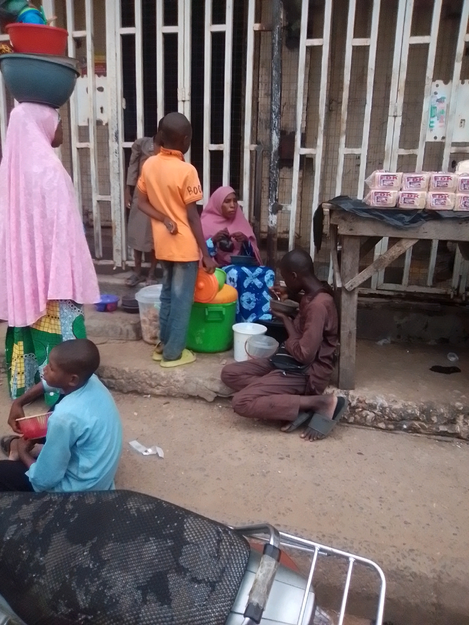 In Arawa, Nigeria, a scene unfolds on a dusty street in front of a building with metal grates. Several people are present. A woman wearing a pink hijab and a colorful patterned skirt carries large bowls stacked on her head. To her right, a young boy in an orange t-shirt and blue jeans stands near a green plastic tub filled with items. A woman in a pink headscarf and blue patterned wrapper sits to the right of the boy. Further to the right, a young person in a brown outfit sits on the ground next to a wooden stall displaying packaged bread. Another young boy in a light blue t-shirt is seated on the ground in the foreground, holding a red bowl. The environment suggests a market or street-side vendor area with various containers and goods scattered around.