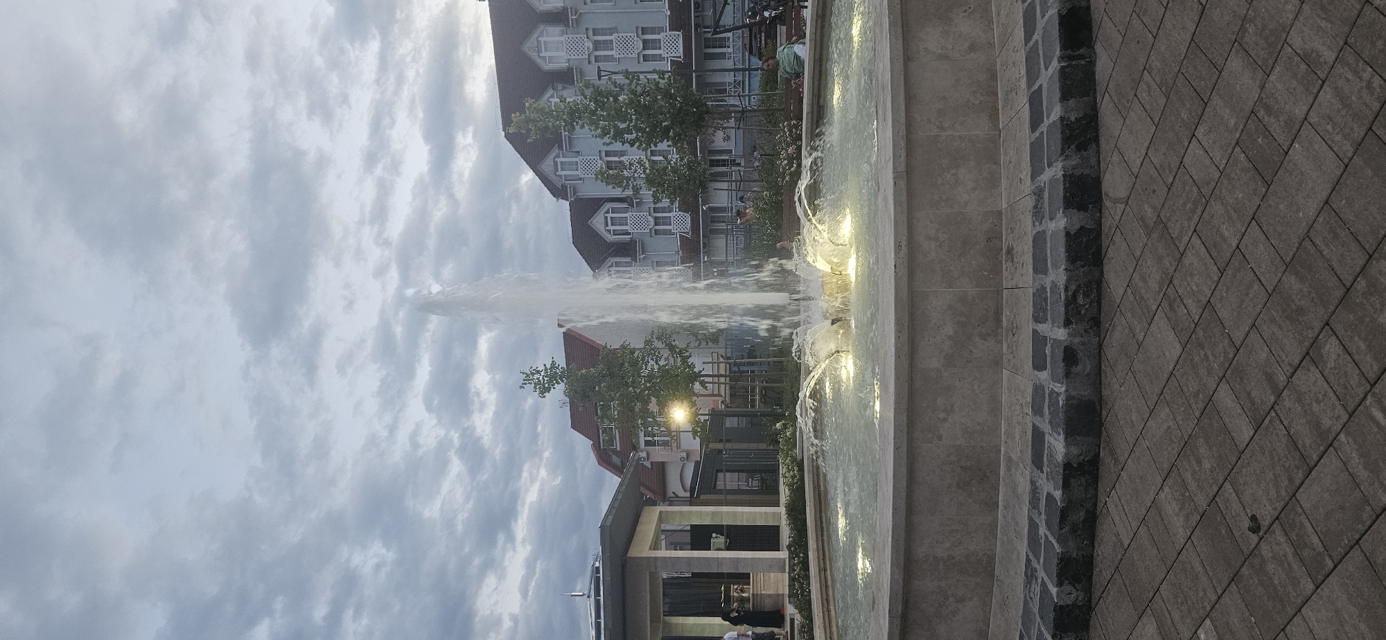 The image shows a fountain in an outdoor urban setting, likely a public square or pedestrian area in Hévíz, Hungary. The fountain is the primary object, with water jets creating a dynamic display. Several buildings with European architectural styles are visible in the background, featuring multiple stories, windows, and decorative elements. Trees are planted around the fountain and between the buildings, adding greenery to the scene. Paved walkways, made of stone or brick, surround the fountain area.
There are a few people visible in the image, some standing near the fountain and others in the background. One person is sitting on a bench by the fountain. The activity taking place is simply the operation of the fountain, with people enjoying the public space.
The time of day appears to be either late afternoon or early evening, as indicated by the lighting from the fountain and a streetlamp, and the slightly overcast sky. The weather seems to be cloudy or partly cloudy.
Notable details include the illuminated water in the fountain, which adds a warm glow to the scene. The contrast between the stone of the fountain and the paved walkway is also apparent. The buildings in the background suggest a developed urban environment.
There is no visible text in the image.
