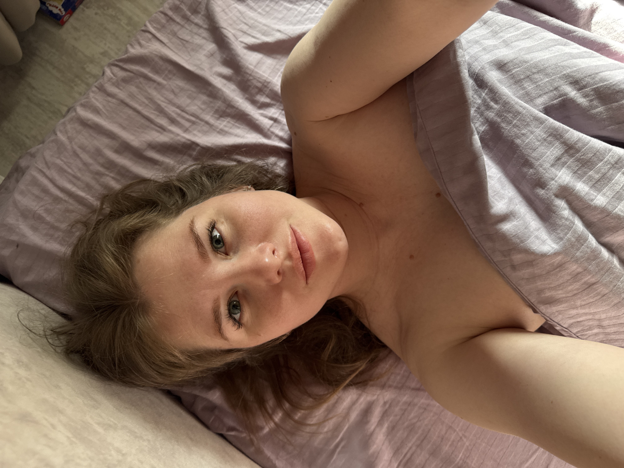 An overhead shot captures a woman lying on her side on a bed, looking directly at the camera. Her head rests on a light-colored surface, possibly a pillow or mattress, with her body partially covered by a light purple, subtly striped sheet or blanket. The bed sheets beneath her are also a similar shade of light purple.
The woman has fair skin, wavy light brown hair, and striking blue-green eyes. Her expression is relaxed, with her lips slightly parted. She is bare-shouldered, with the sheet covering her torso and part of her arm. Sunlight appears to be streaming from the right, illuminating the blanket and her skin with a soft glow, suggesting it is daytime, possibly morning.
In the upper left corner, beyond the bed, a glimpse of a light-colored floor and a small red and white object, possibly a box or bag, is visible. There is no visible text in the image. The scene suggests an intimate moment of repose indoors.