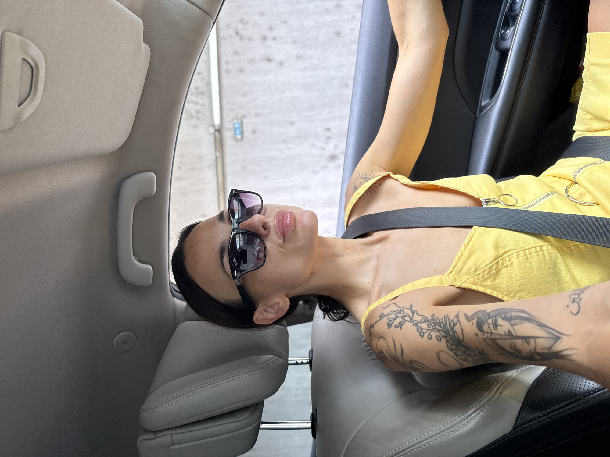 The image depicts a young adult female lying down in the interior of a car, captured from an overhead or slightly upward angle. The woman has dark hair and is wearing black sunglasses, with the brand name "Ray-Ban" visible on the arm. Her lips are slightly parted, adorned with gloss or lipstick. She is dressed in a bright yellow jumpsuit or overall, featuring silver zipper details and rings, and a dark seatbelt is fastened across her midsection. Both of her arms and shoulders are covered in tattoos, including a detailed, intricate design on her left arm and a symbol, possibly "Om," on her right shoulder.
The car's interior is visible, featuring a beige sun visor, a grab handle, and what appears to be a small speaker grille on the ceiling panel. The seats are made of light and dark-colored leather or faux leather. A car door panel with controls and a handle is also visible. The background outside the car is blurred, showing a light-colored, textured surface that suggests a concrete structure, possibly indicating a parking garage or an urban setting during the daytime. The woman appears to be posing for a photograph in a relaxed manner. The location context for this scene is Pleven, Bulgaria.