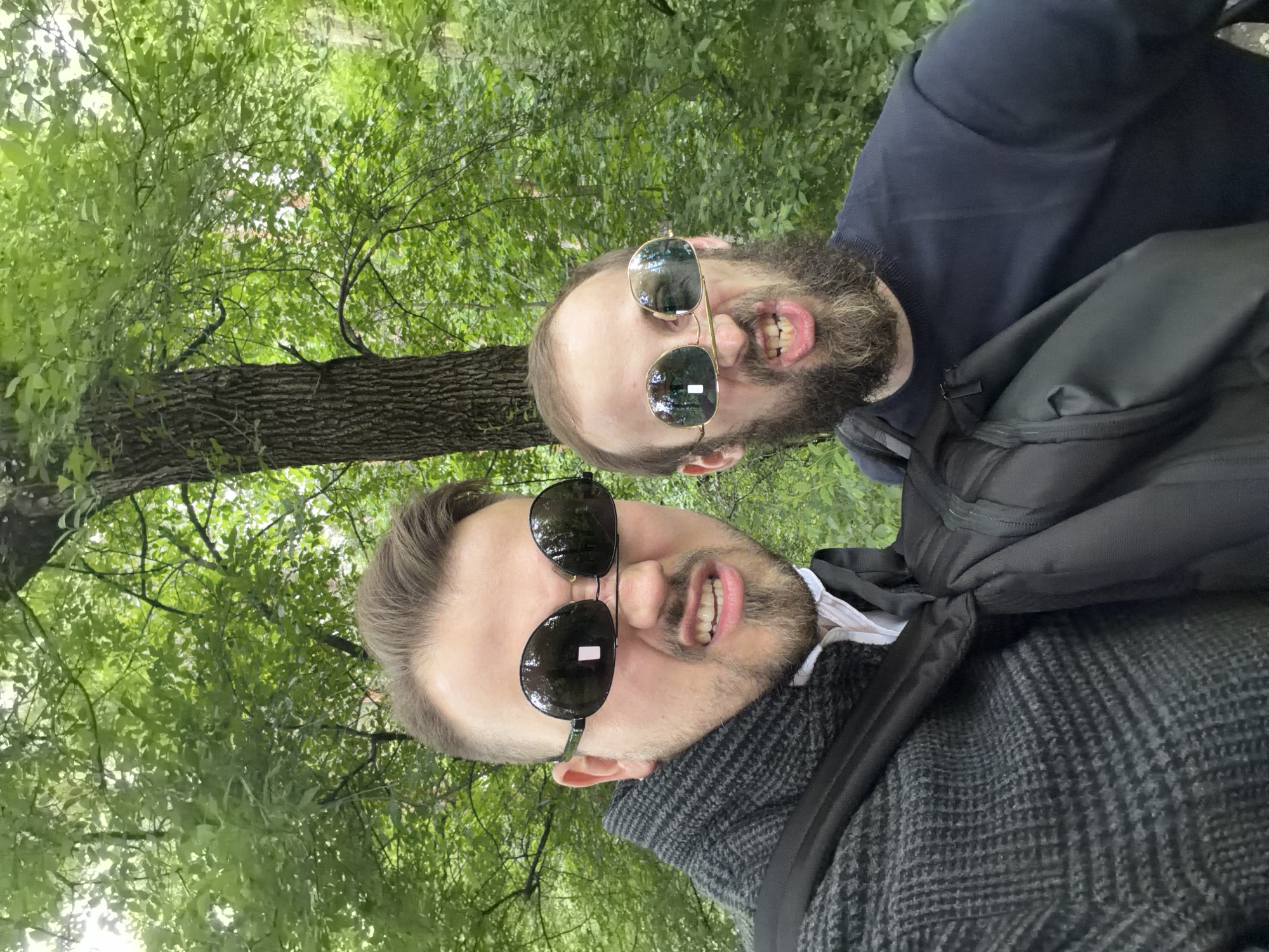 Two men are pictured from a low angle, possibly a selfie, against a backdrop of green foliage, likely trees in a park setting. The location appears to be Moscow, Russia, as indicated by the architectural style or cultural elements visible.
The man in the foreground has dark hair and a beard, wearing a dark sweater and sunglasses, and a black backpack strap is visible across his shoulder. His expression is exaggerated with a toothy grin. The second man, positioned behind and slightly above the first, also wears sunglasses and has a beard. He appears to be wearing a dark blue sweater. Both men are making exaggerated facial expressions. The background consists of dense greenery, suggesting a wooded or park area. The overall impression is casual and candid, likely taken during an outdoor excursion.