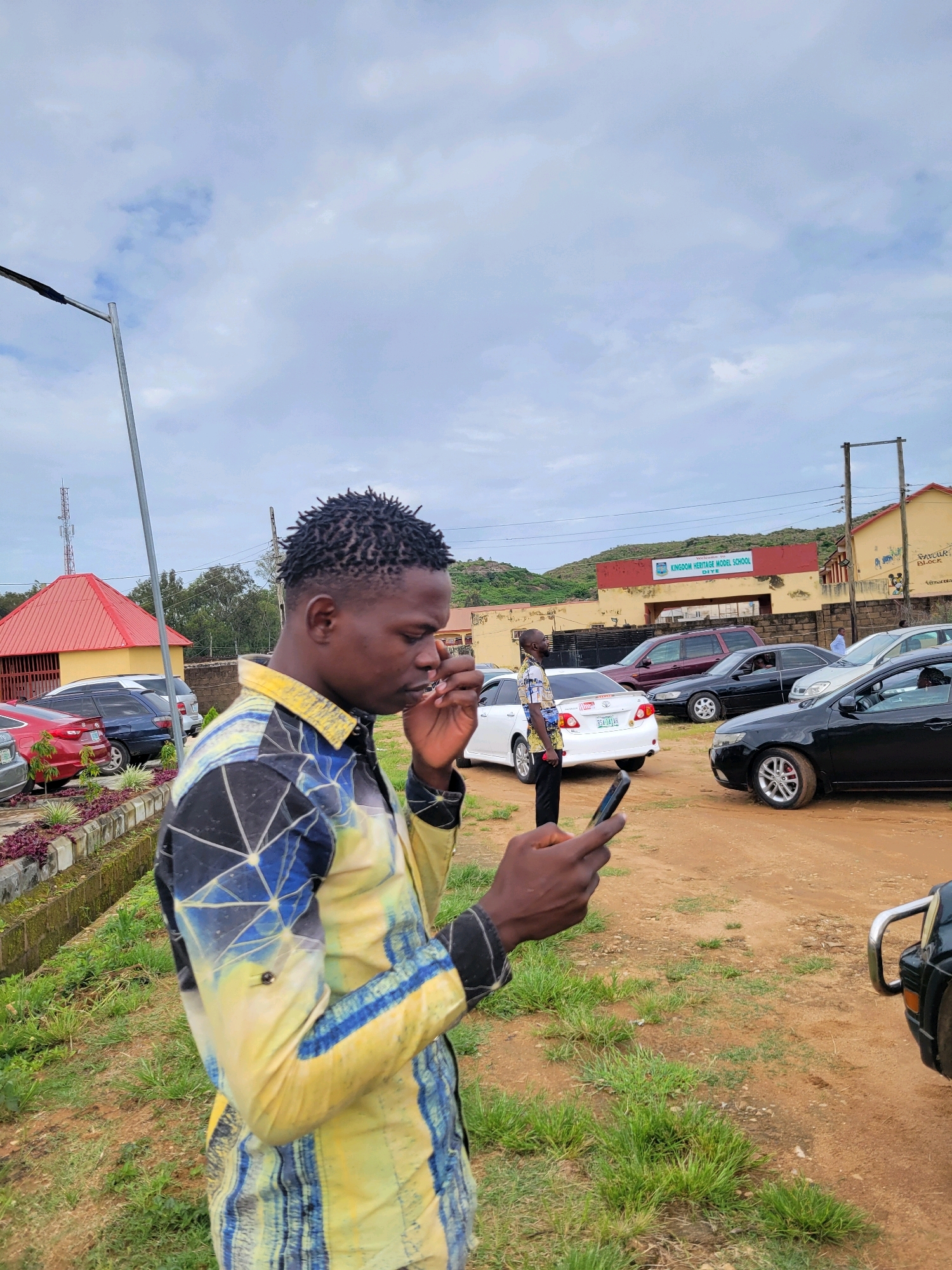 In the city of Gura Top, Nigeria, an individual with dark, braided hair stands outdoors, holding a mobile phone in their right hand while their left hand is near their face. They are wearing a long-sleeved, collared shirt with a vibrant geometric pattern in yellow, blue, and black. The background shows a dirt road with several parked cars, including a black sedan in the foreground and a white sedan and a maroon SUV further back. Behind the vehicles is a low-rise building with a red-tiled roof and a yellow facade, possibly part of the "Kingdom Heritage Model School" as indicated by a sign on the building. There are also trees and a hilly landscape visible in the distance under a cloudy sky. A lamppost is positioned to the left of the frame.