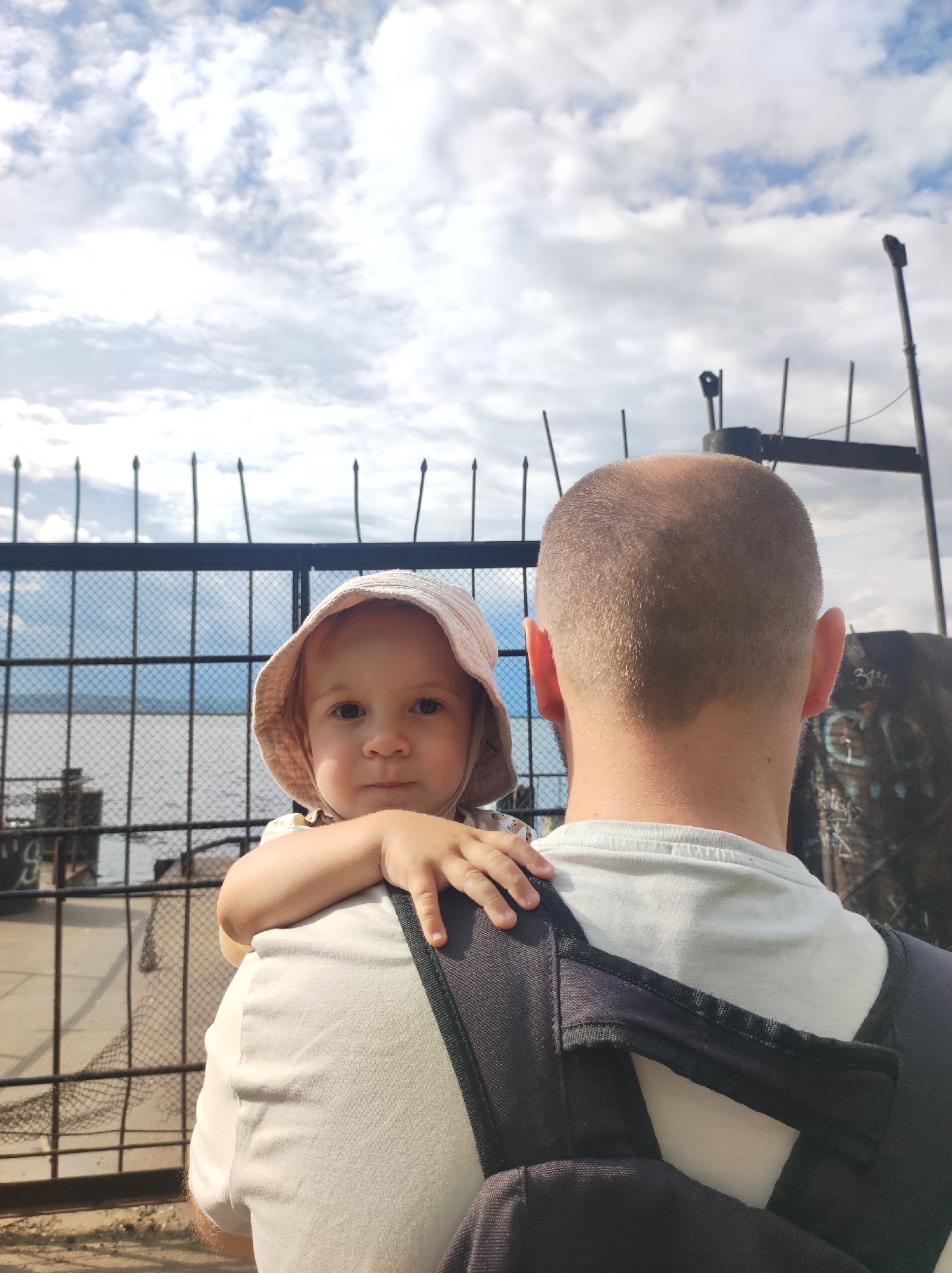 An over-the-shoulder, medium shot captures a toddler being carried by an adult on a cloudy day, near a body of water. The toddler, facing the camera, wears a light-colored sun hat and has their right arm resting on the adult's shoulder, their hand gently placed on the adult's neck. The adult, seen from behind and mostly out of frame, has a shaved head and is wearing a light-colored t-shirt and a dark backpack. In the background, a black metal fence with pointed finials separates the foreground from a body of water, possibly the Kama River, which is visible behind the fence. The sky is filled with white and gray clouds. The overall scene suggests a casual outing in Chaikovsky, Russia, likely near a waterfront area or embankment.