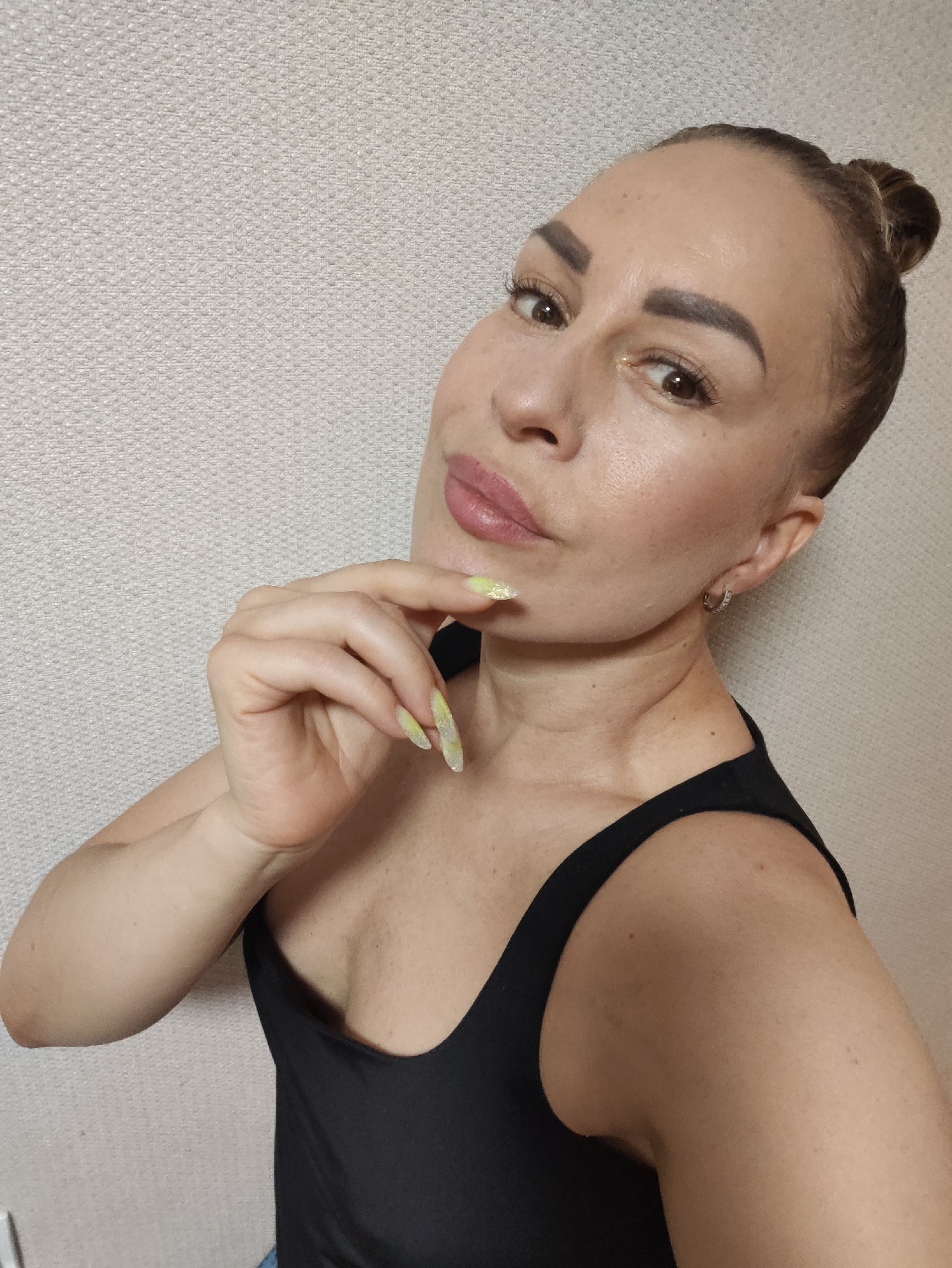 A medium shot shows a woman with her hair pulled up in a bun, looking at the camera with one hand on her chin. She is wearing a black tank top and has long, light green fingernails with glitter. The background is a plain, textured wall. There is no indication of the city of Almaty, Kazakhstan, or any specific location within a city.