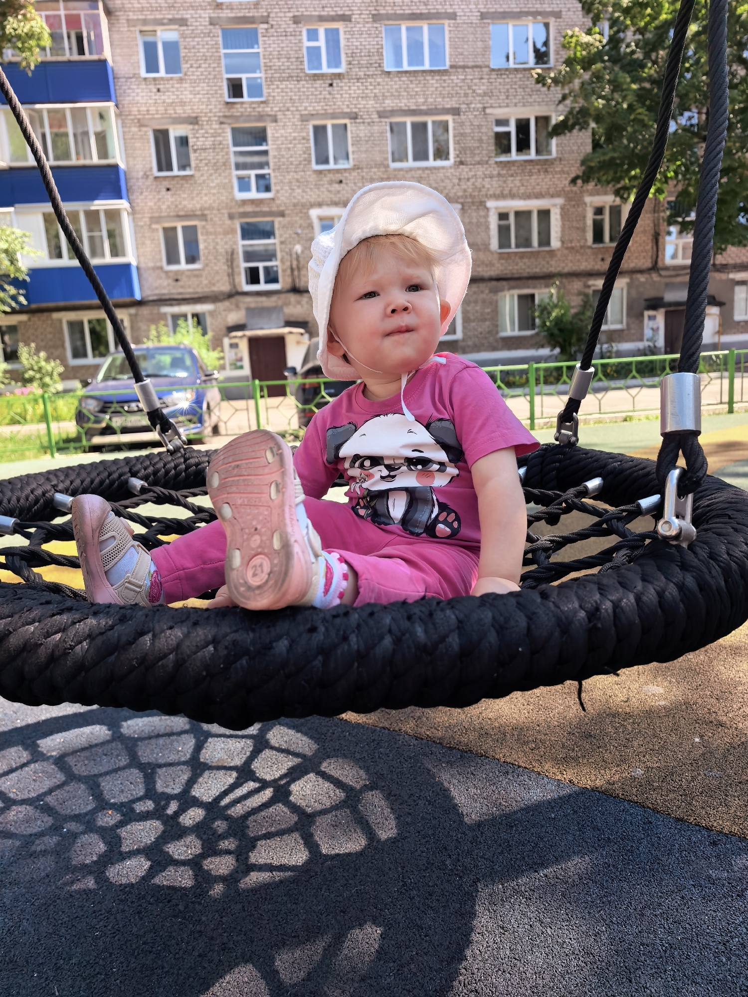 A young child, appearing to be approximately 1-2 years old, sits on a large, black rope swing designed for playground use. The child is wearing a pink t-shirt with a panda graphic and pink pants, along with a white sun hat and pink shoes. The swing is suspended by thick black ropes attached to metal fixtures, and its shadow is cast on the rubberized playground surface below. In the background, a multi-story brick apartment building with numerous windows is visible, along with trees and a blue car parked in front of the building. The setting appears to be a residential courtyard or playground area in Sterlitamak, Russia, under daylight conditions.