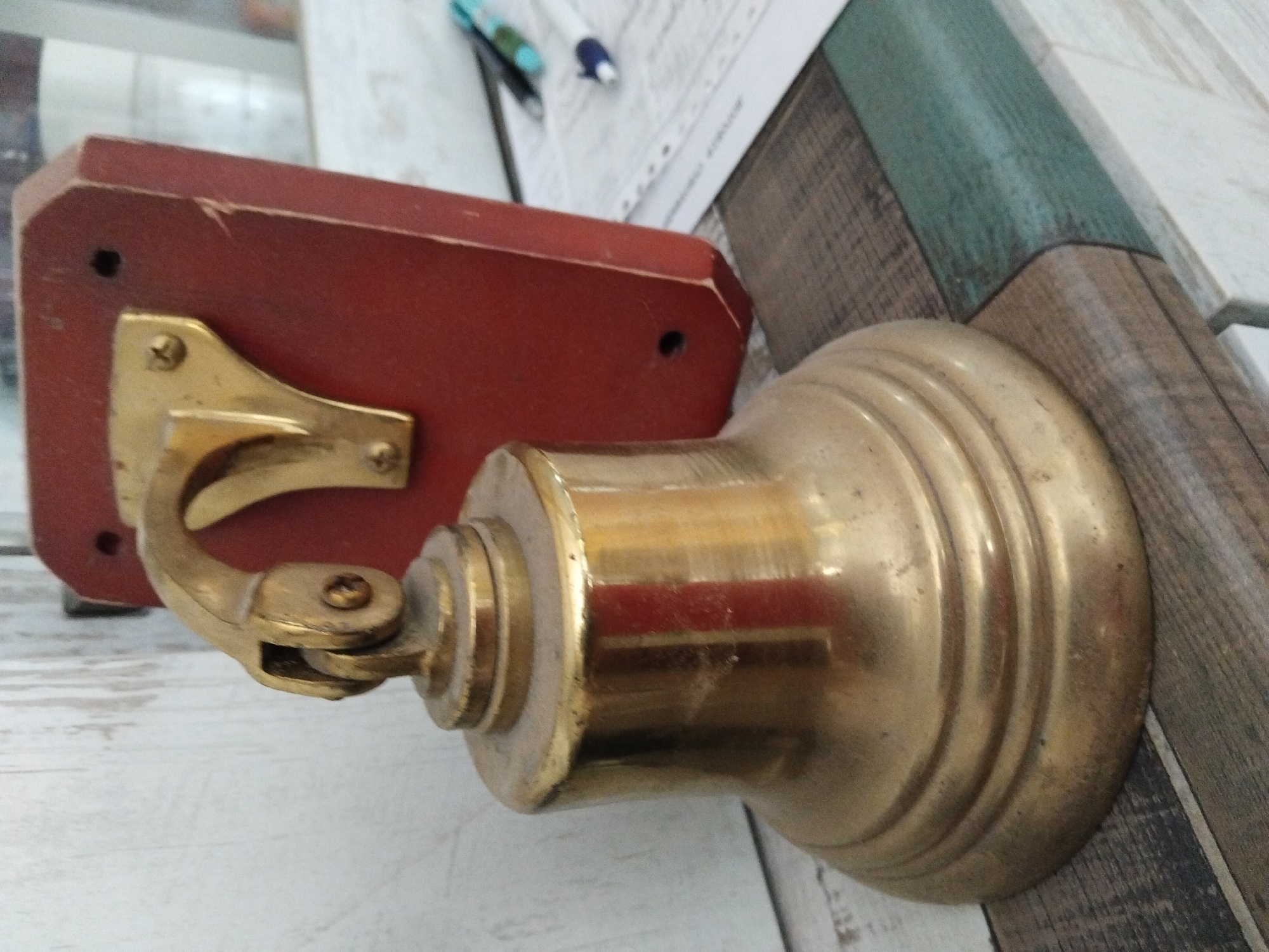 A vintage brass bell is mounted on a red wooden plaque. The bell is bell-shaped with a flared base and decorative concentric rings around the lower portion. It hangs from a brass bracket attached to the plaque. The plaque has a visible screw holding the bracket and appears to be mounted on a vertical surface. In the background, there is a wooden wall or partition, and a blurred area with papers and pens, suggesting an indoor setting. The overall impression is one of an old or decorative item, possibly for signaling or as an antique. The image does not contain any information about the city of Kyiv, Ukraine.