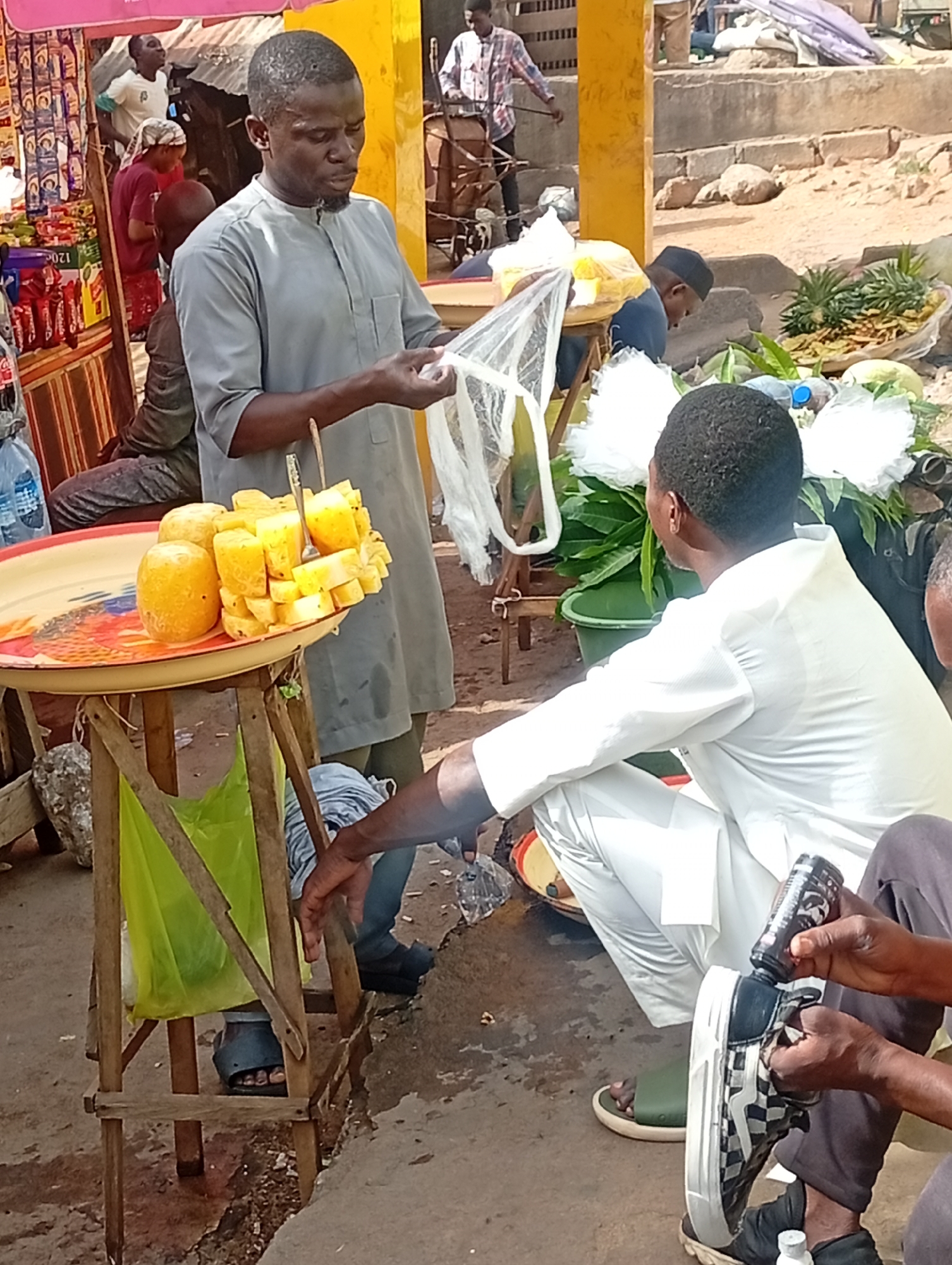 The image depicts a street scene in Maiburiji, Nigeria. A man in a gray tunic is holding open a plastic bag, presumably for a customer. He stands next to a small wooden table holding a tray of cut pineapples. A man in white clothing is seated on the ground nearby. Another man is visible in the lower right corner, cleaning a checkerboard-patterned shoe with black shoe polish. Additional figures and market stalls are visible in the background, along with produce such as pineapples and cabbages. The setting appears to be an outdoor market area with a mix of vendors and customers.