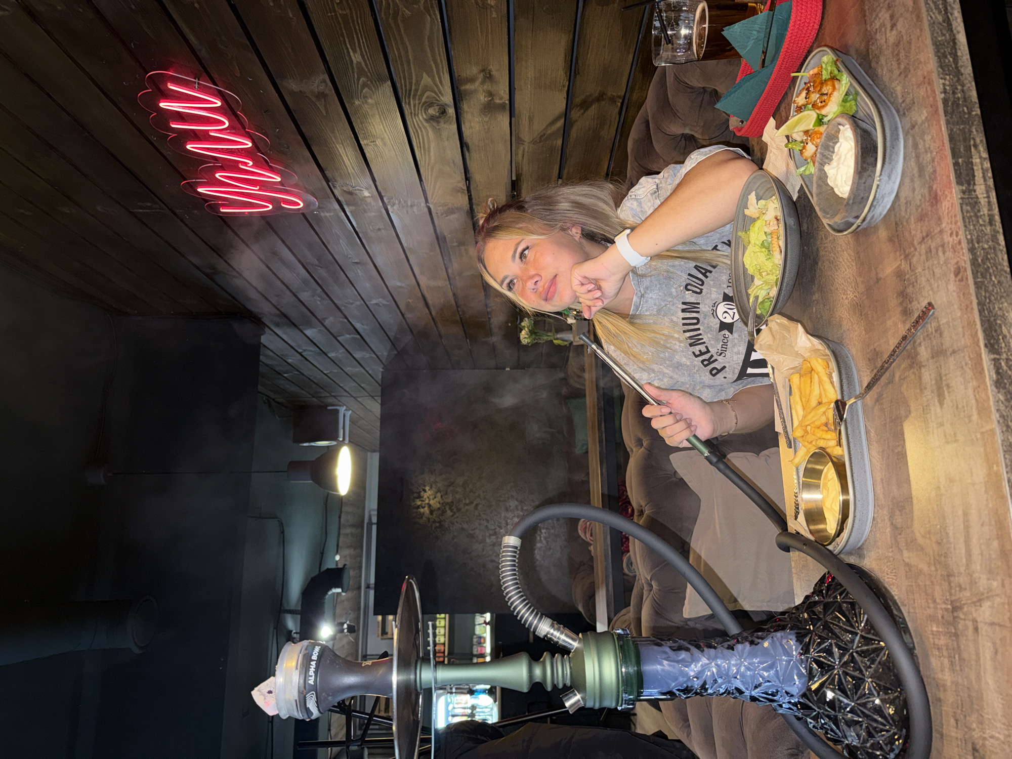 The image depicts a relaxed indoor scene, likely in a lounge or restaurant in Chaikovsky, Russia, during the evening or night. A young woman with long blonde hair, wearing a grey t-shirt, is seated at a dark wooden table, holding the hose of a hookah. She gazes thoughtfully upwards and to her left, with a calm expression.
On the table, an assortment of food and drinks is present. These include a plate of fried shrimp garnished with lime and accompanied by a white dipping sauce, a bowl of green salad, and a separate dish of french fries with another small container of sauce. A clear glass with a beverage and straw, along with a red woven basket, are also visible. The prominent hookah features a dark, patterned base and a green-toned stem. The hookah bowl bears the text "ALPHA BOWL".
The establishment has a warm, somewhat dim atmosphere. The ceiling is constructed from dark wooden panels. A distinctive red neon sign, spelling out "МЯТА" (MYATA, meaning 'Mint' in Russian), is affixed to the wooden ceiling. In the background, industrial-style wall lights illuminate parts of the room, and the woman appears to be seated on a plush, upholstered sofa. Wisps of smoke, likely from the hookah, are visible in the air. The text on the woman's t-shirt reads "PREMIUM QUALITY Since 20".